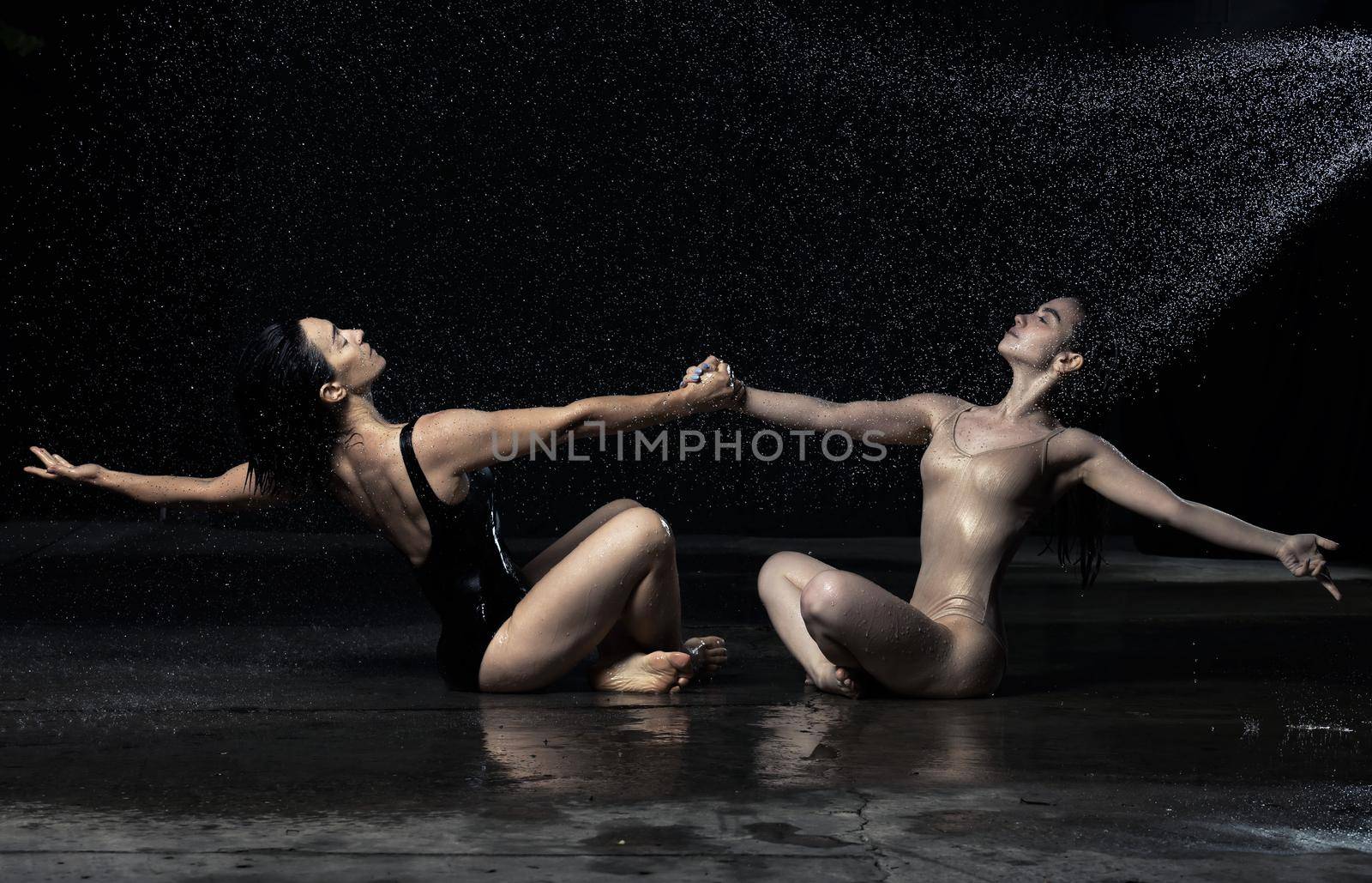 two beautiful women of Caucasian appearance with dark hair are sitting on the asphalt in drops of water on a black background, holding hands and looking up. People are wearing bodysuits