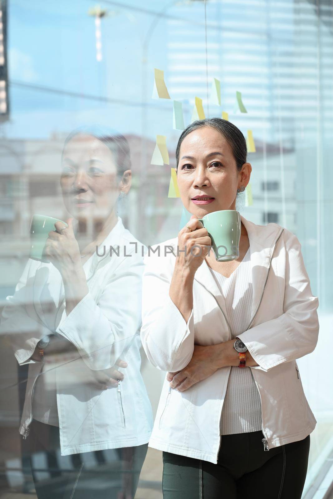 Elegant senior female business leader holding coffee cup and standing near large window at office with city buildings in background.