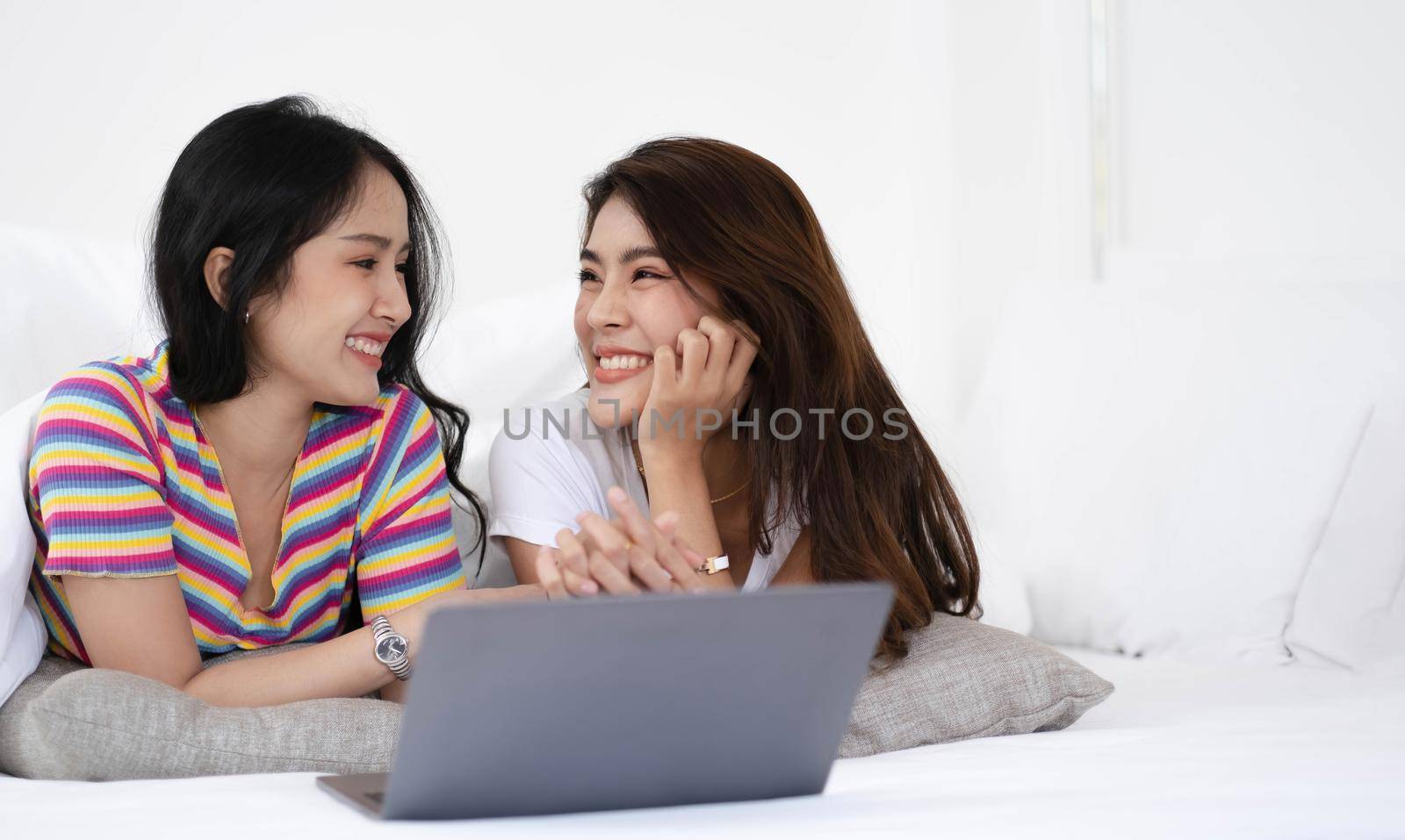 Young asian LGBT Lesbian couples are using laptops to search for information to buy a new home. while embracing each other in bed. by wichayada