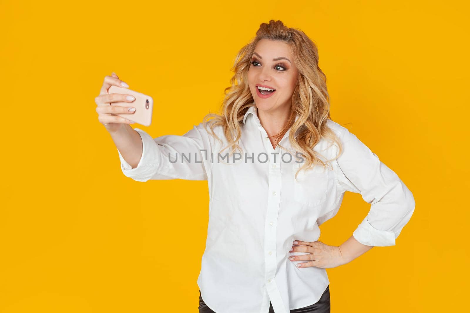 A blond woman in a white middle-aged shirt takes a selfie on a smartphone, holds a mobile phone in her outstretched hand and tries to look good in a photo on a yellow background.