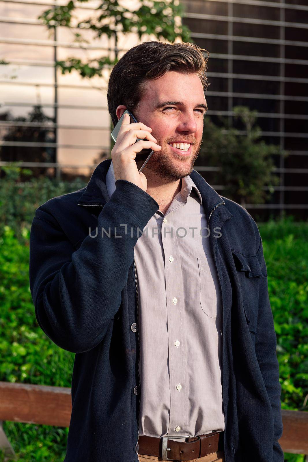 smiling caucasian business man talking by phone in a park next to an office building, concept of technology and communication