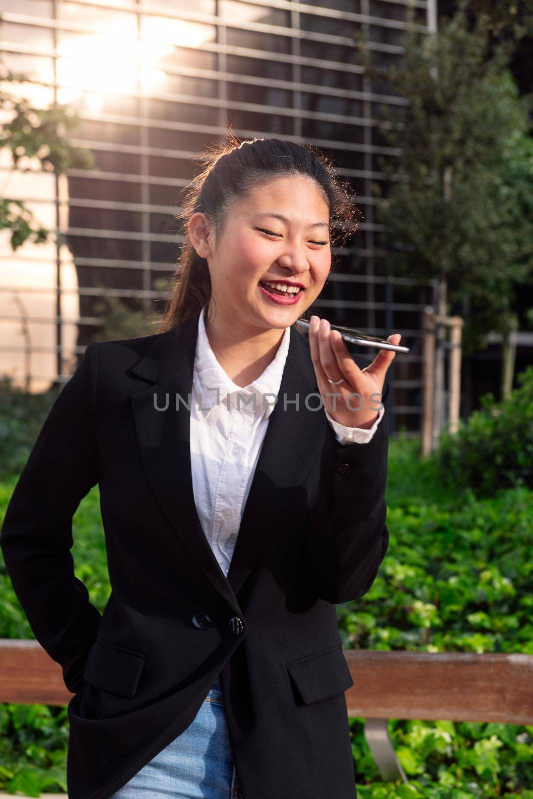 smiling asiatic business woman talking by phone in a park next to an office building, concept of technology and communication