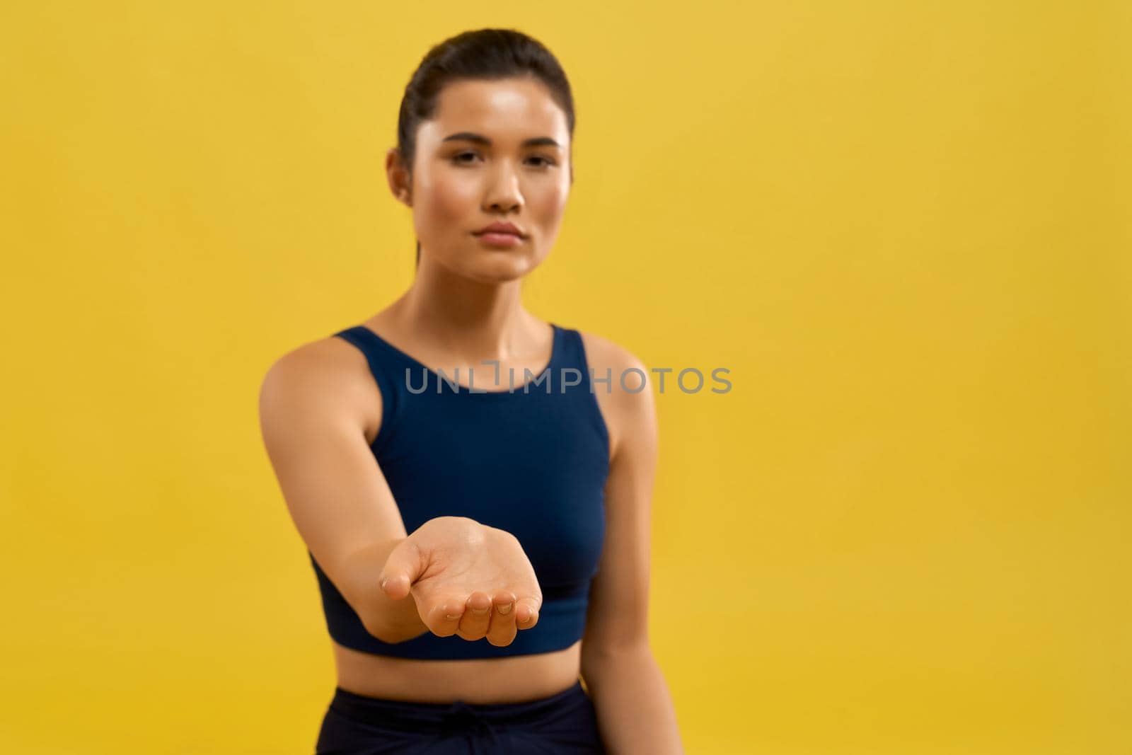 Front view of sporty girl standing, raising hand forward, looking at camere, begging. Brunette with pony tail stretching, yoga doing. Isolated on yellow studio background.