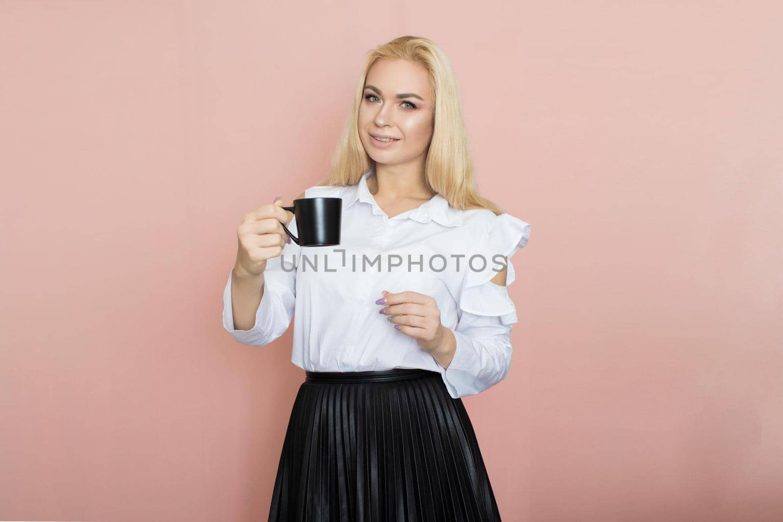 Beauty, fashion portrait. Elegant business style. Portrait of a beautiful blonde woman in white blouse and black skirt posing at studio on a pink background. Holding black cup in her hands. Drinking coffee, tea