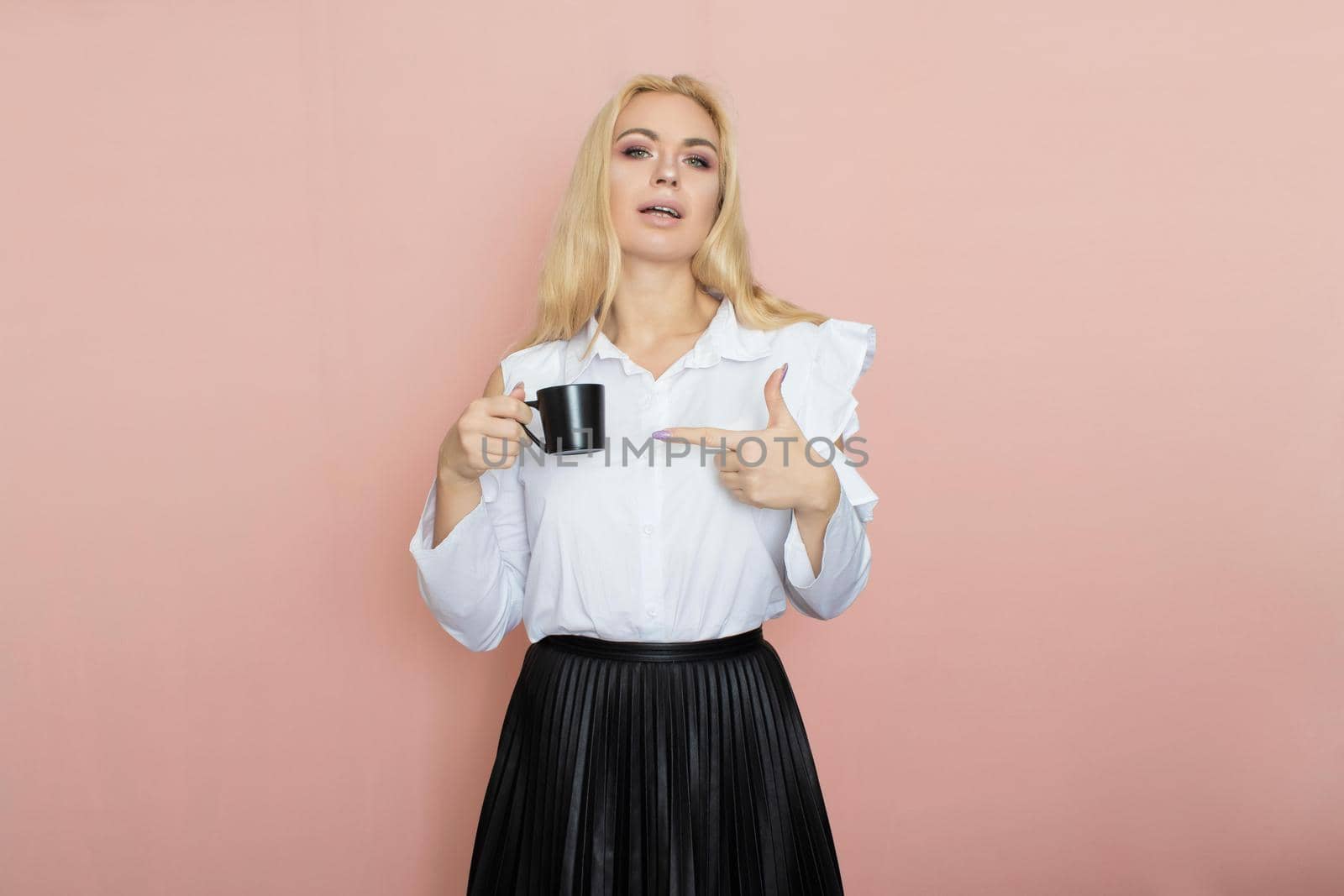 Beauty, fashion portrait. Elegant business style. Portrait of a beautiful blonde woman in white blouse and black skirt posing at studio on a pink background. Holding black cup in her hands. Drinking coffee, tea