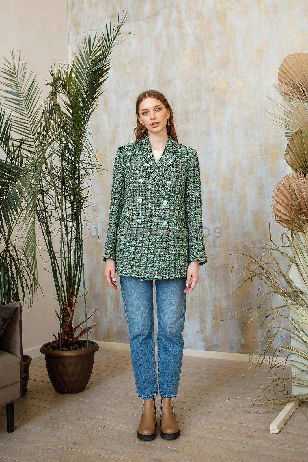 A girl in a green fashionable jacket and blue jeans on a studio background with plants.