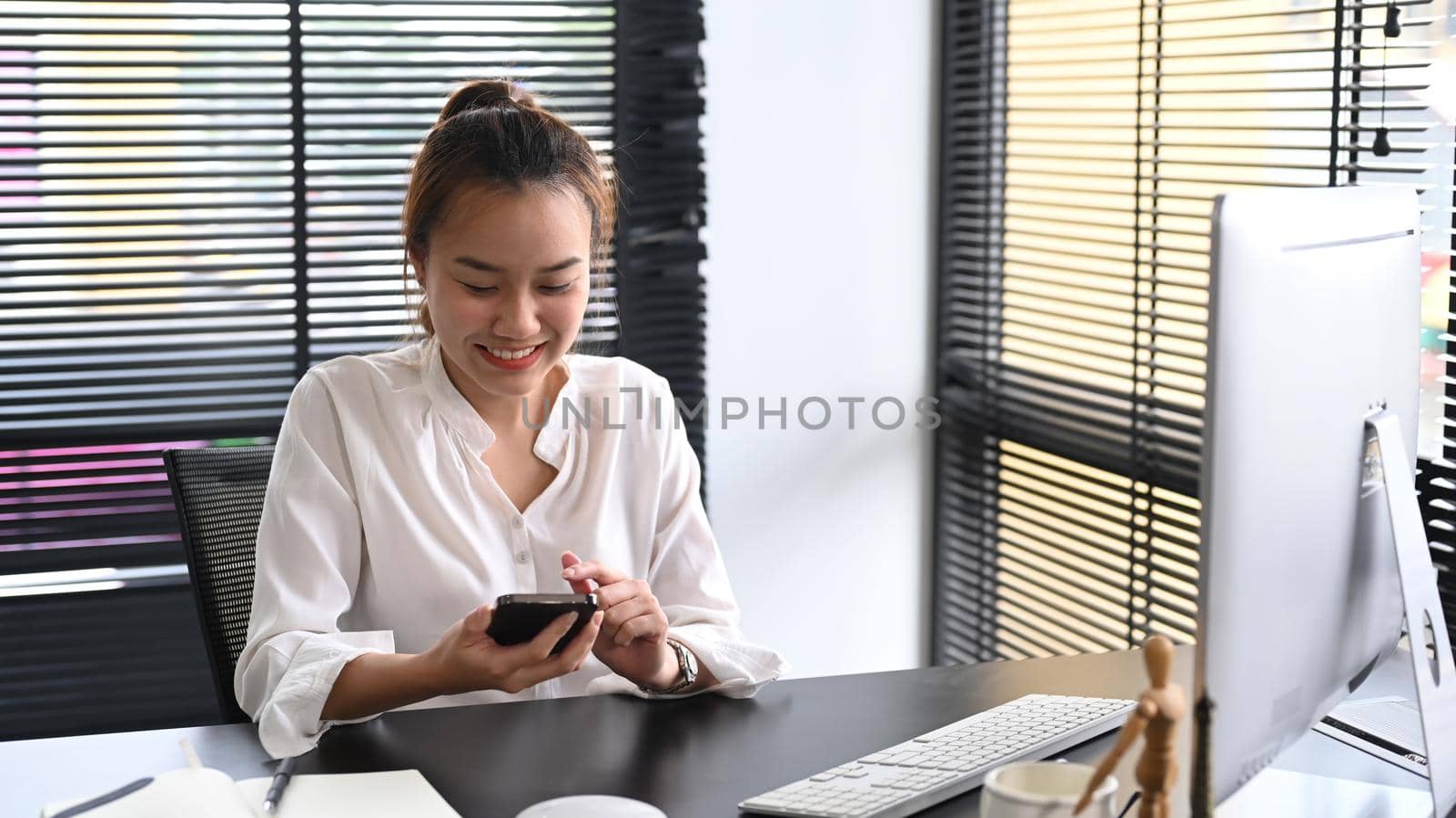 Happy millennial asian woman sitting at her workplace and using mobile phone for communicating in social network.