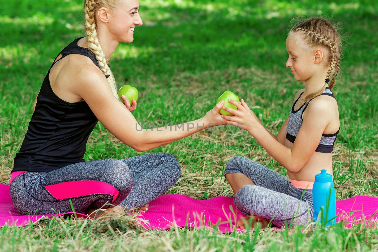 Mother and her daughter in sportswear holding a green apples and sitting on the roll mat in park.