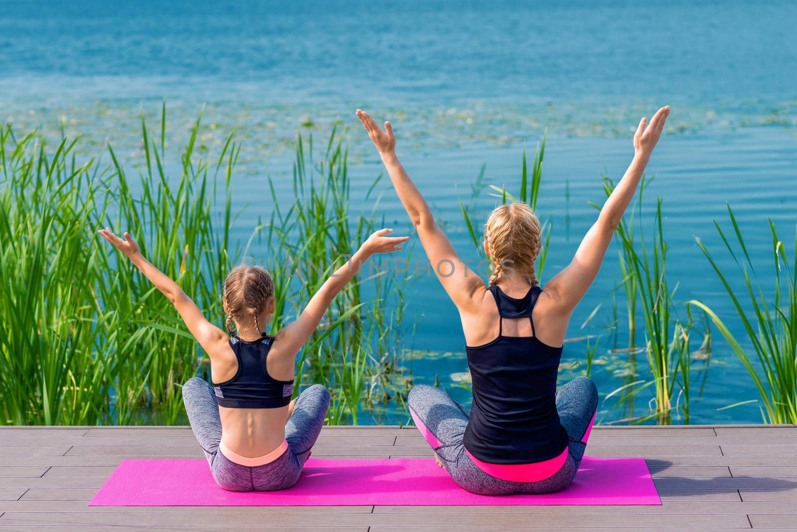 Mother and daughter doing yoga at the shore of the lake in summer.