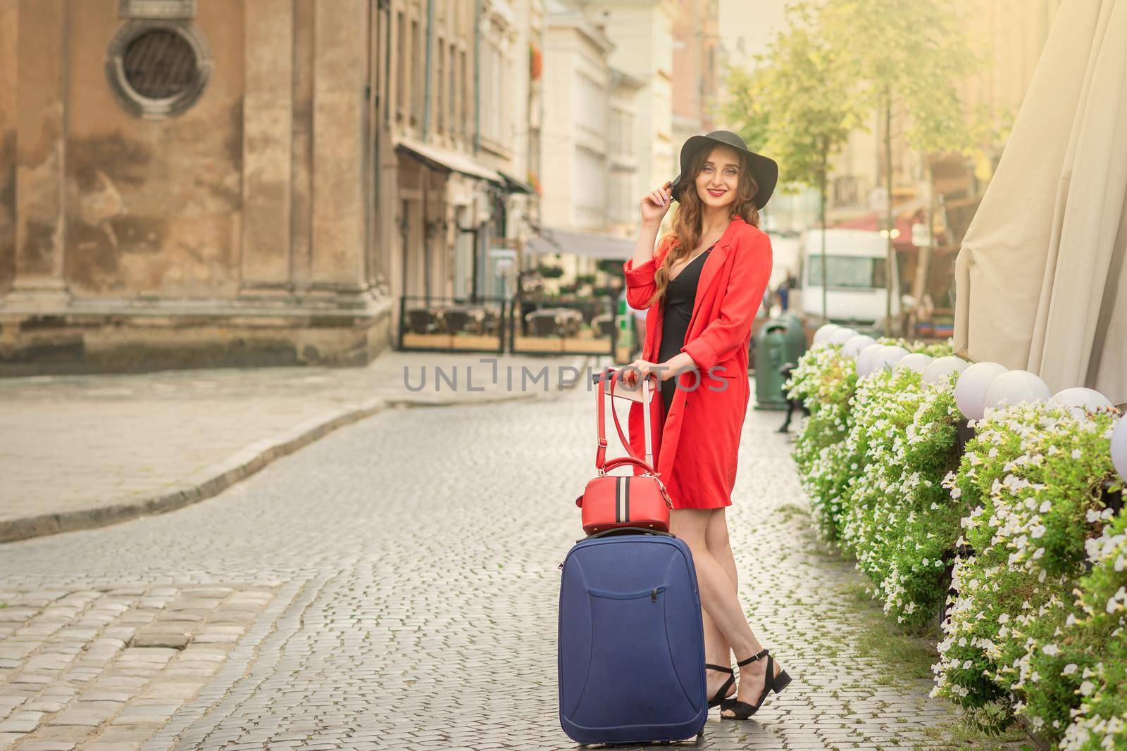 Cheerful white woman standing on the old street wearing hat with suitcase smiling at camera on background of city.