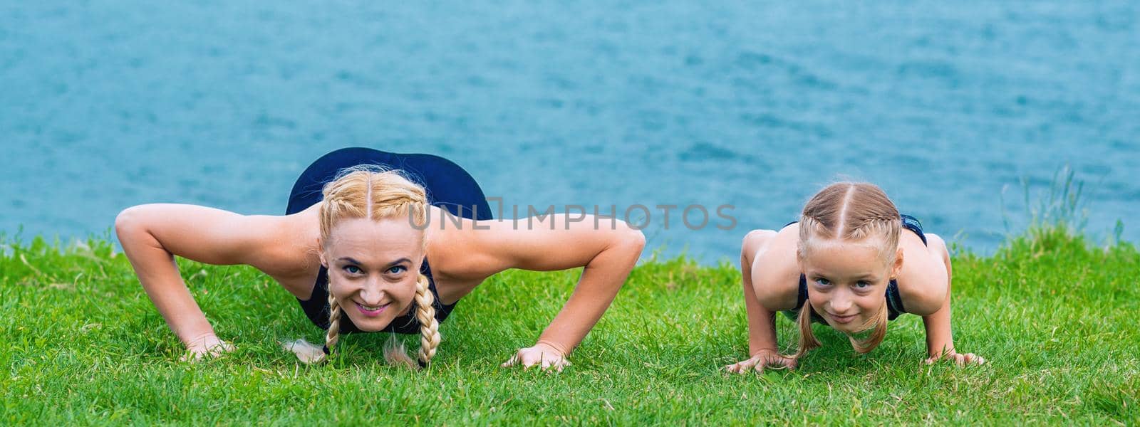 Little girt and woman are doing exercises on the grass at the shore of the lake.