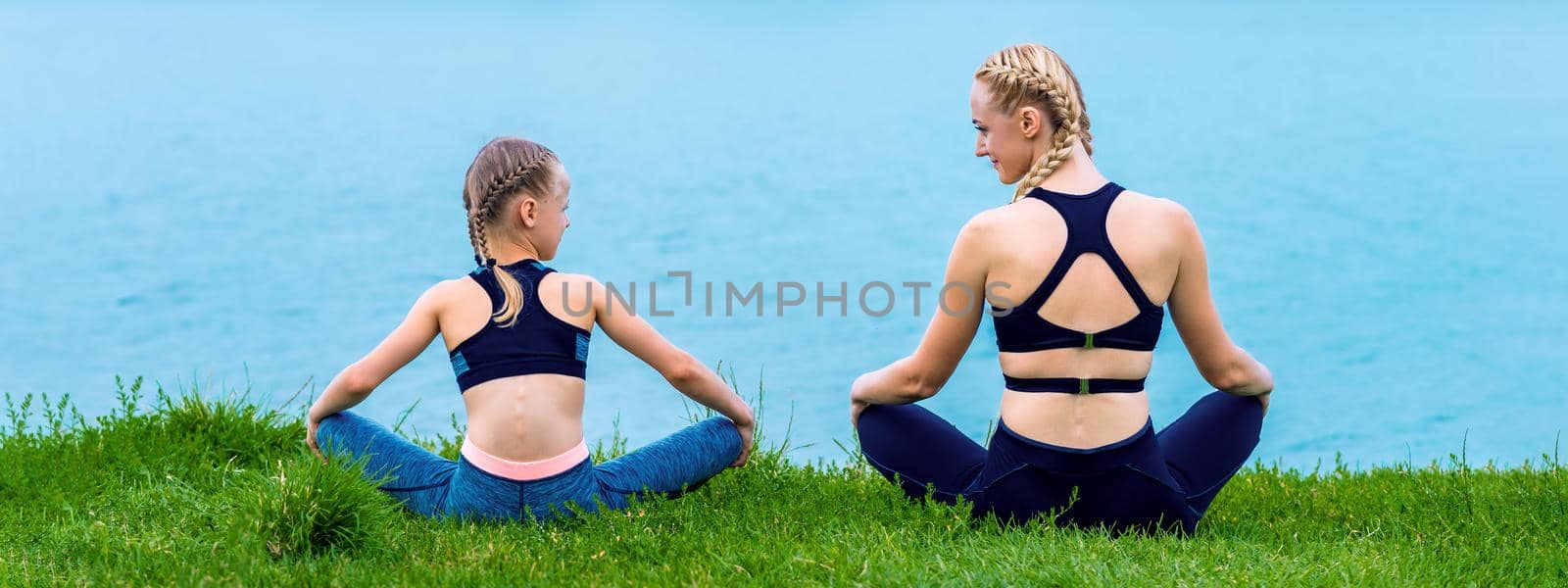 Mother and daughter doing yoga exercises on grass of the shore of the lake.