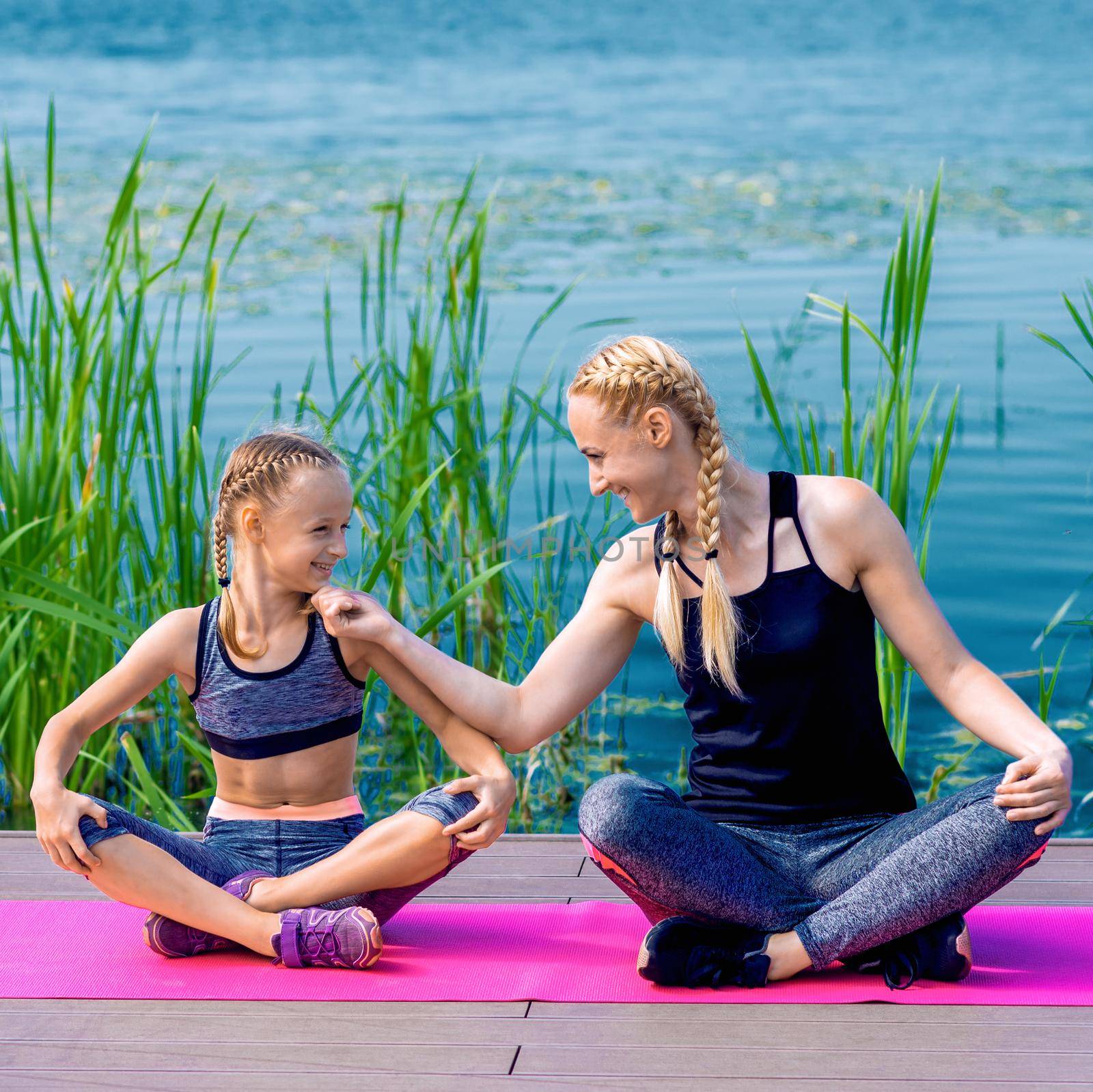 Portrait of mother and daughter sitting on yoga mat by the lake outdoors. Healthy and exercise concept.
