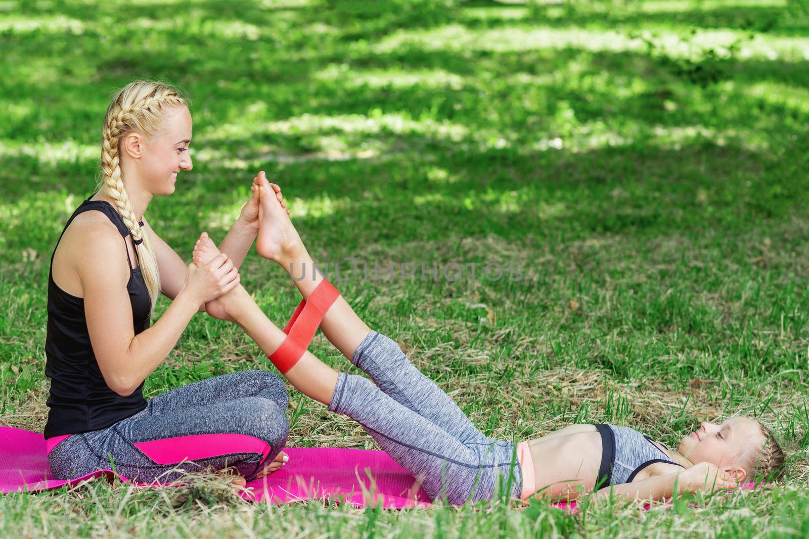 Young woman is training little girl's legs by fitness gum on mat in the park.