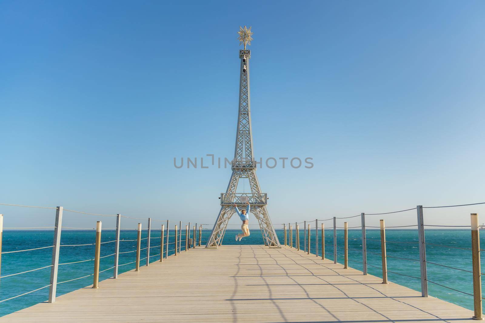 Large model of the Eiffel Tower on the beach. A woman walks along the pier towards the tower, wearing a blue jacket and white jeans. by Matiunina