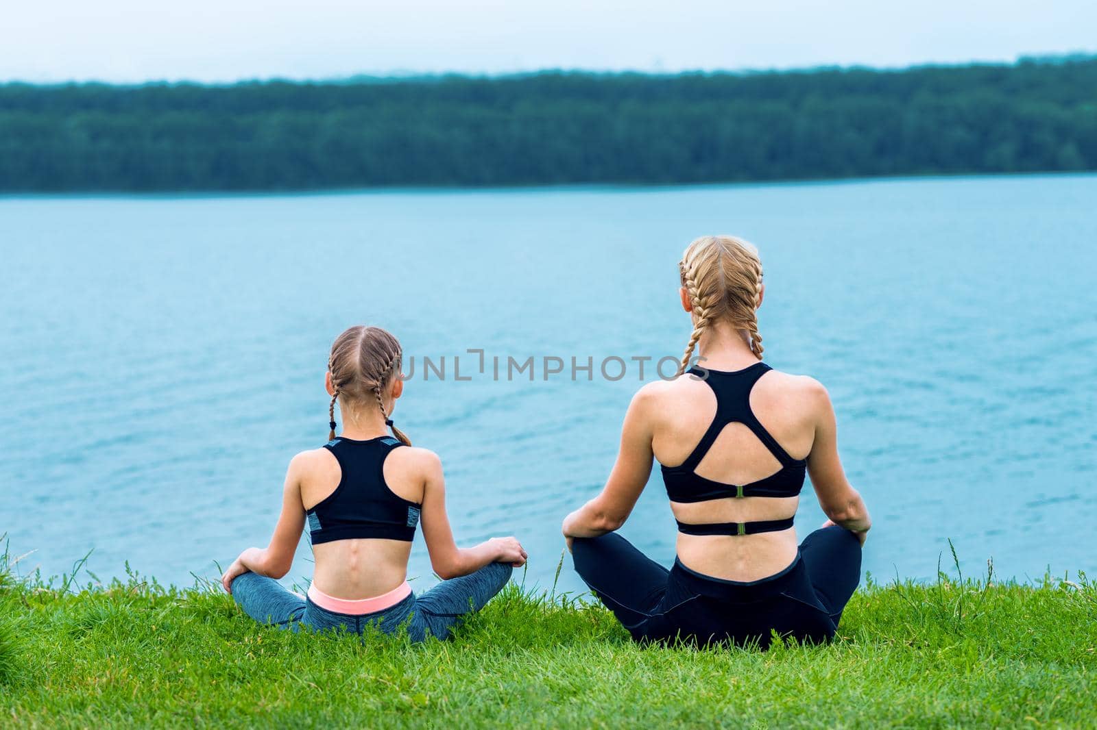 Mother and daughter doing yoga exercises on grass of the shore of the lake.