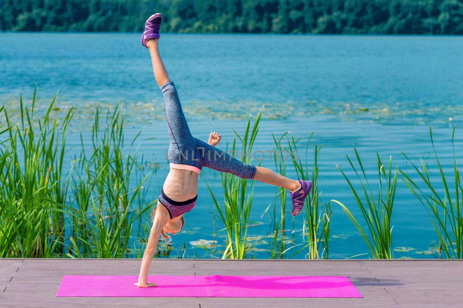 Cute girl is doing acrobatic handstand on roll mat by the lake.