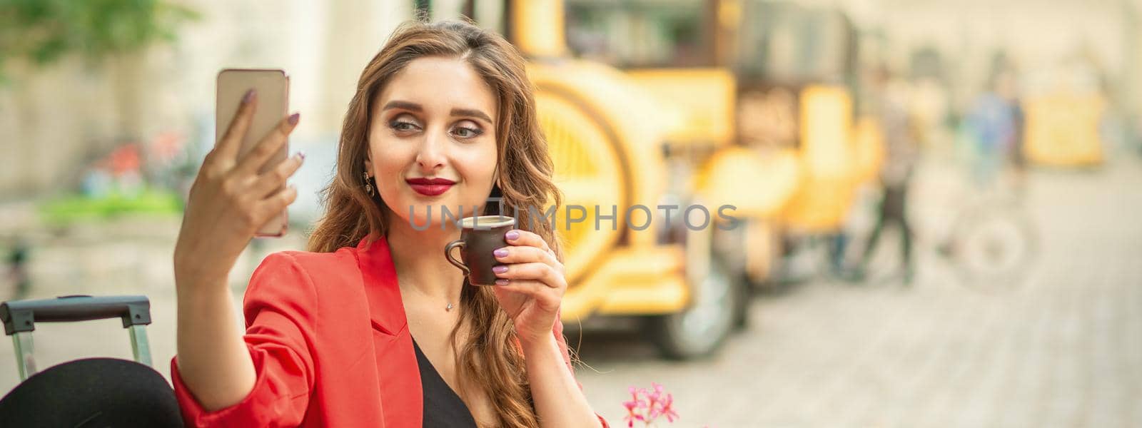 Beautiful brunette woman taking pictures of herself on a cellphone holding cup of coffee at cafe outdoors.