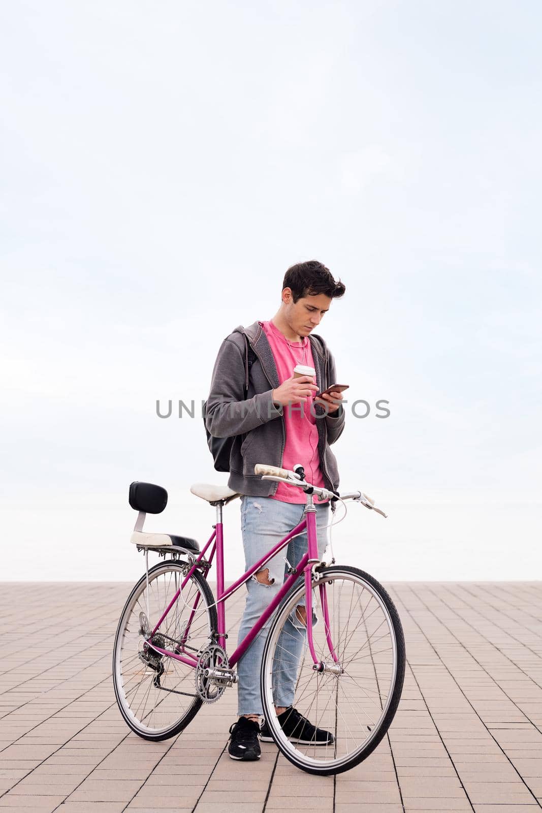 vertical photo of a young man with bicycle holding a coffee while using his mobile phone, concept of communication and sustainable urban transportation, copy space for text