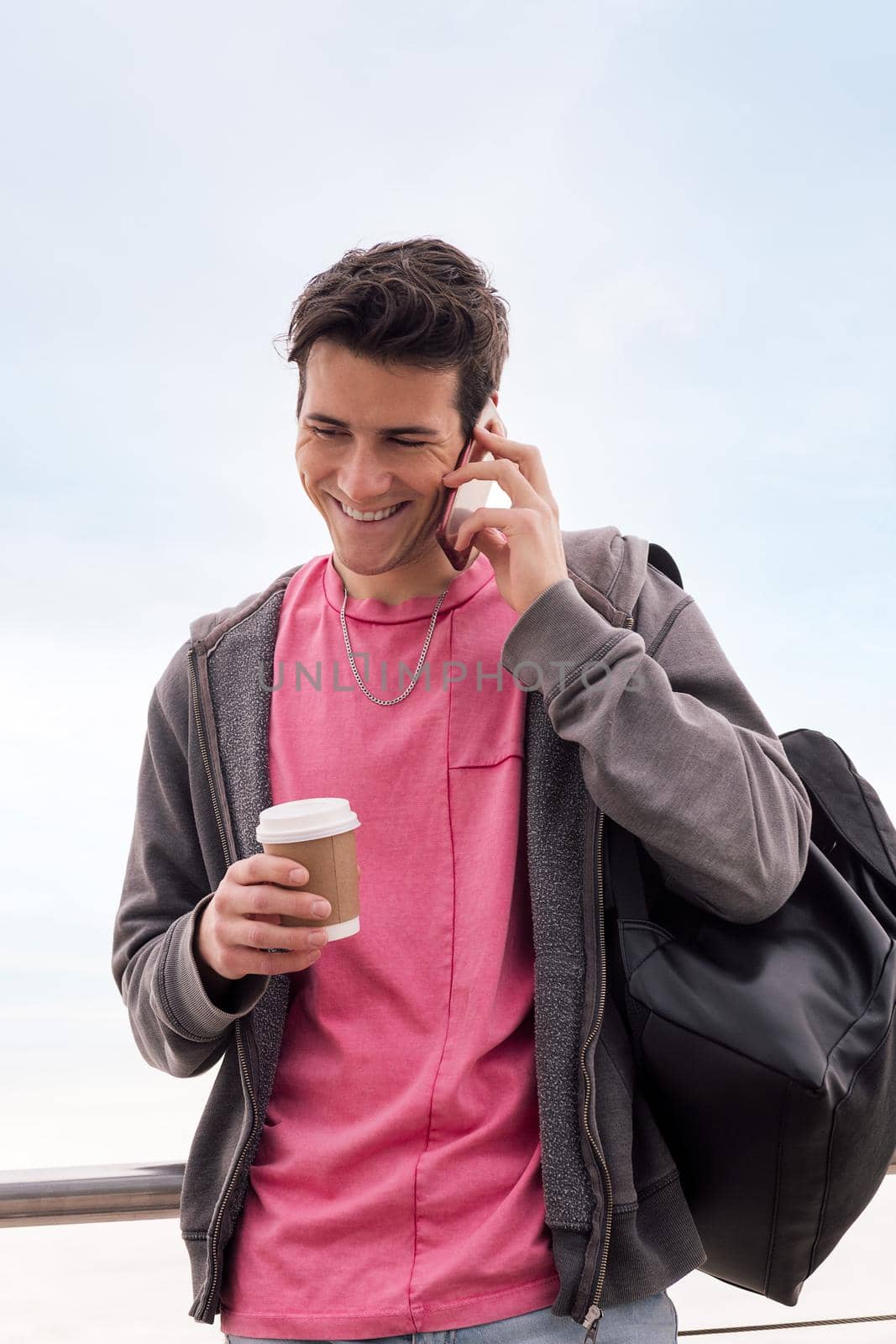 vertical photo of a happy young man with a coffee smiling while talking by mobile phone outdoors, concept of technology of communication and urban lifestyle, copy space for text