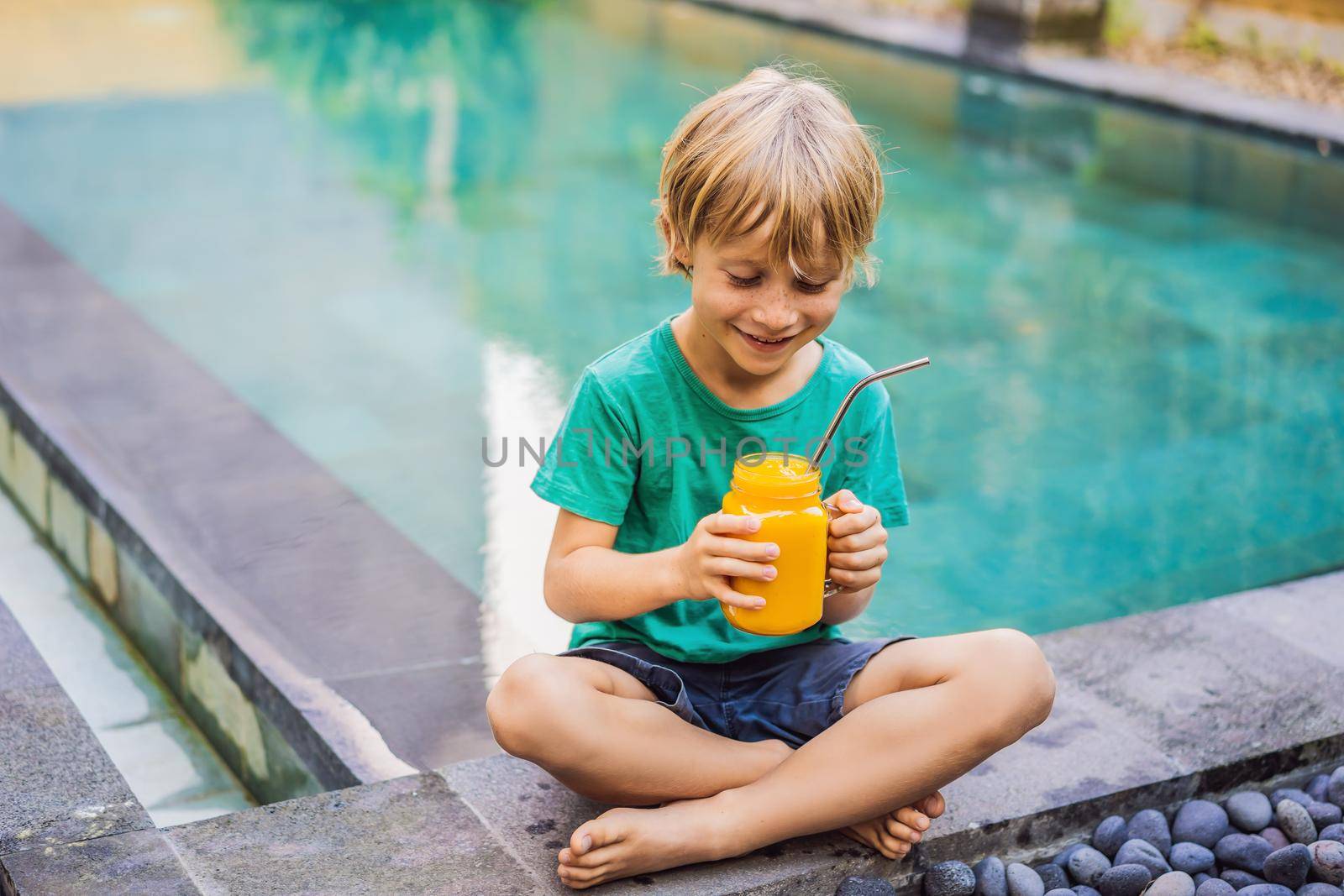 Boy drinking juicy smoothie from mango in glass mason jar with steel straw on the background of the pool. Healthy life concept, copy space.