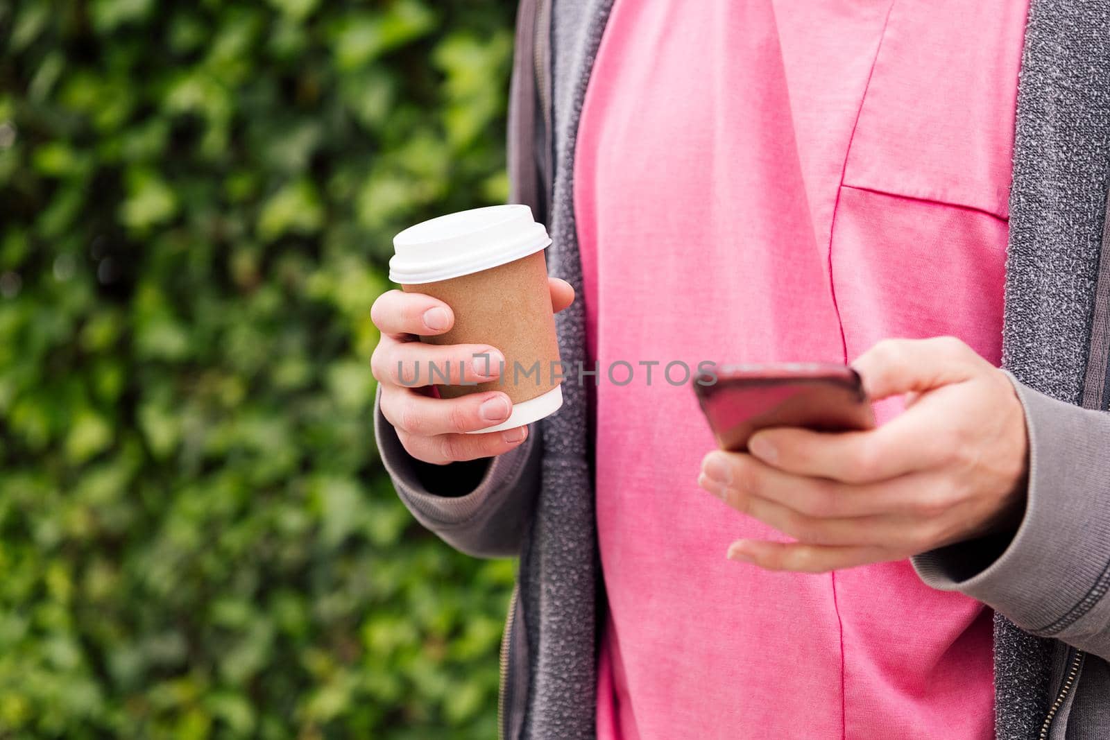 detail of the hands of an unrecognizable young man with a coffee using phone on a green plants background, concept of youth and technology of communication, copy space for text