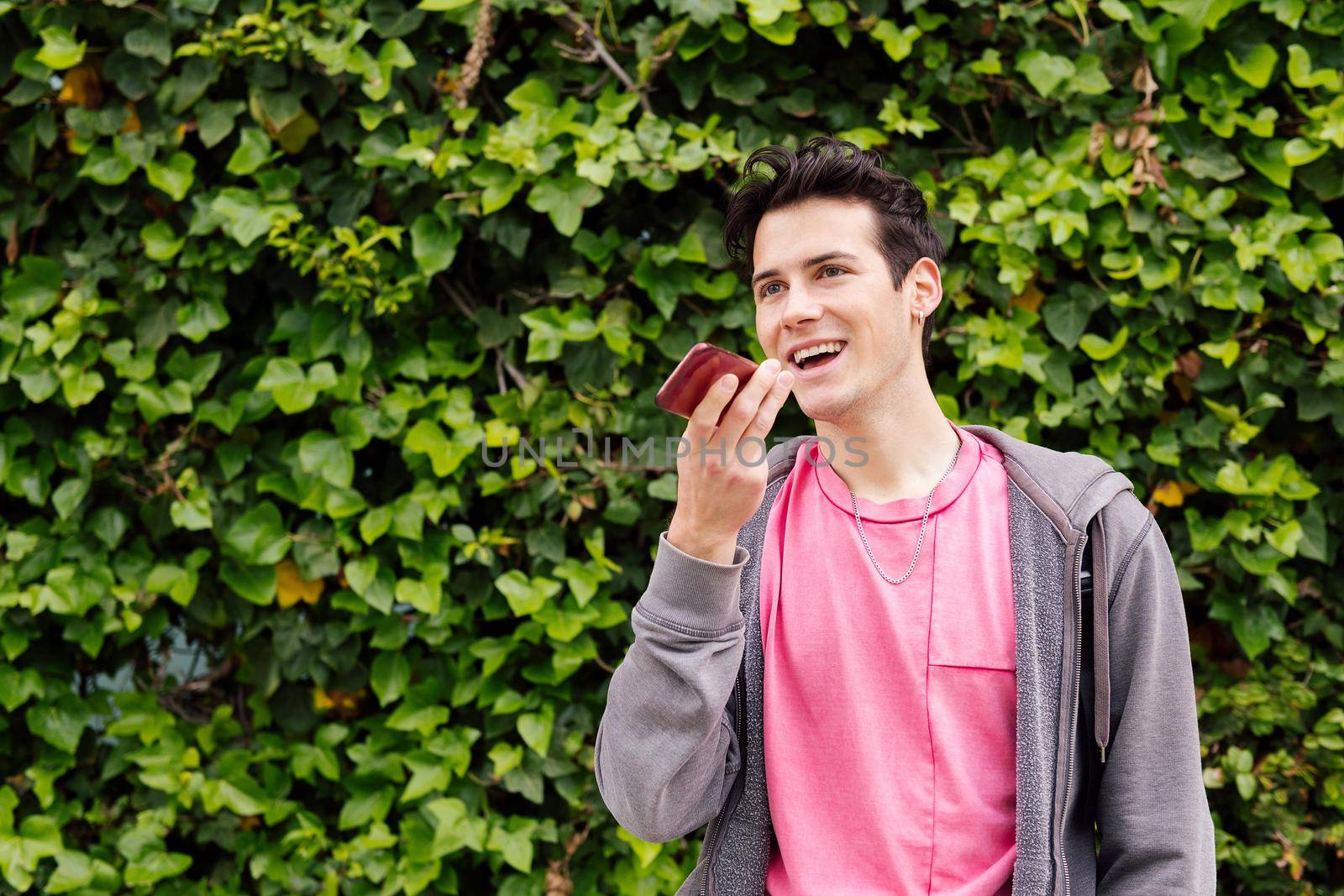 happy teen boy smiling while recording a voice message on mobile phone on a green plants background, concept of youth and technology of communication, copy space for text