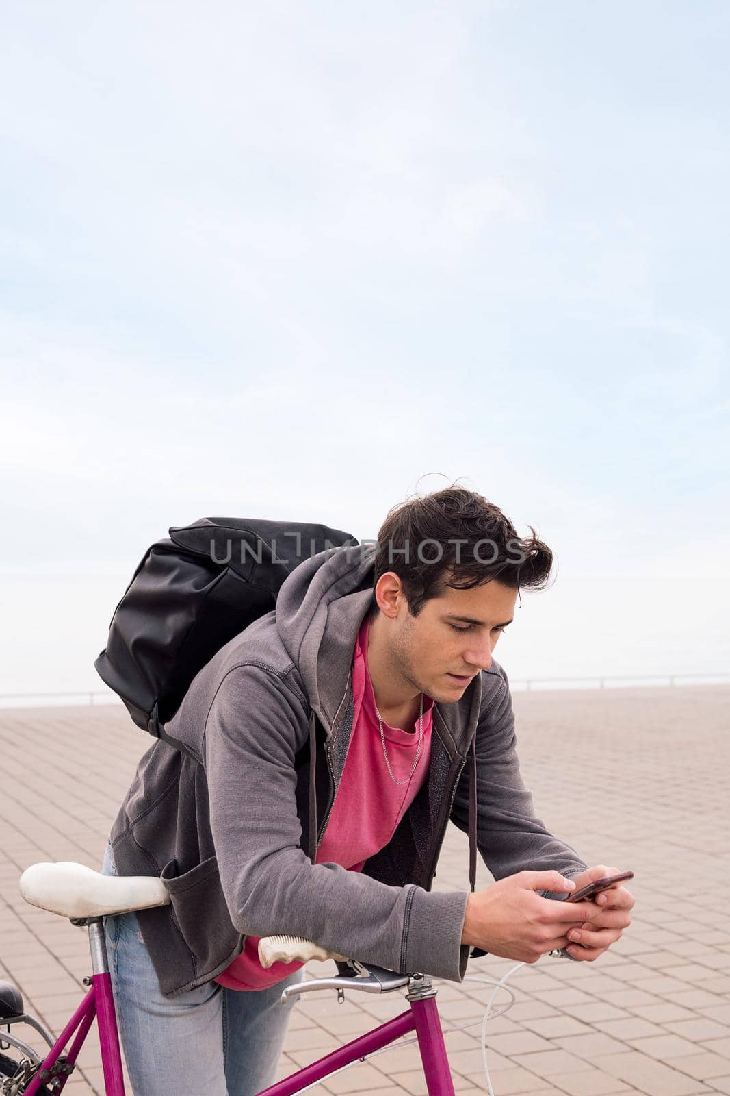 vertical photo of a handsome young student with backpack leaning on a bike using a mobile phone, concept of communication and ecological urban transportation, copy space for text