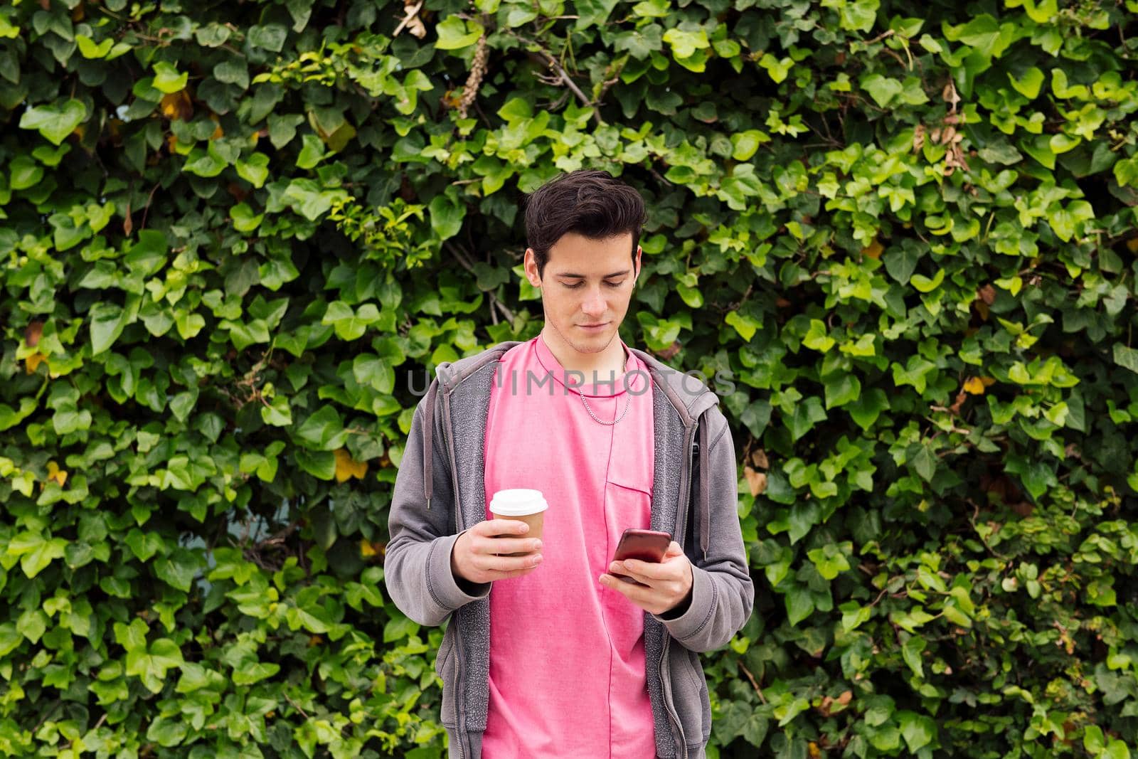young man with a coffee using phone on a green plants background, concept of youth and technology of communication, copy space for text