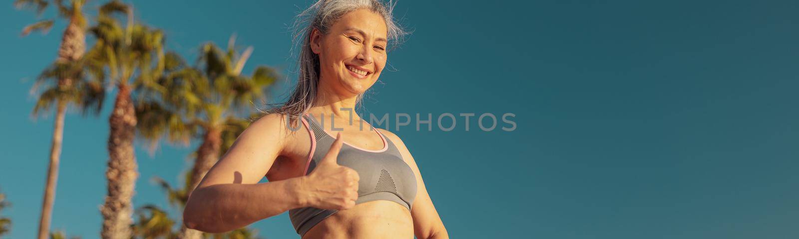 Motivated smiling lady in sports top and leggings resting after training by the seashore and looking at the camera