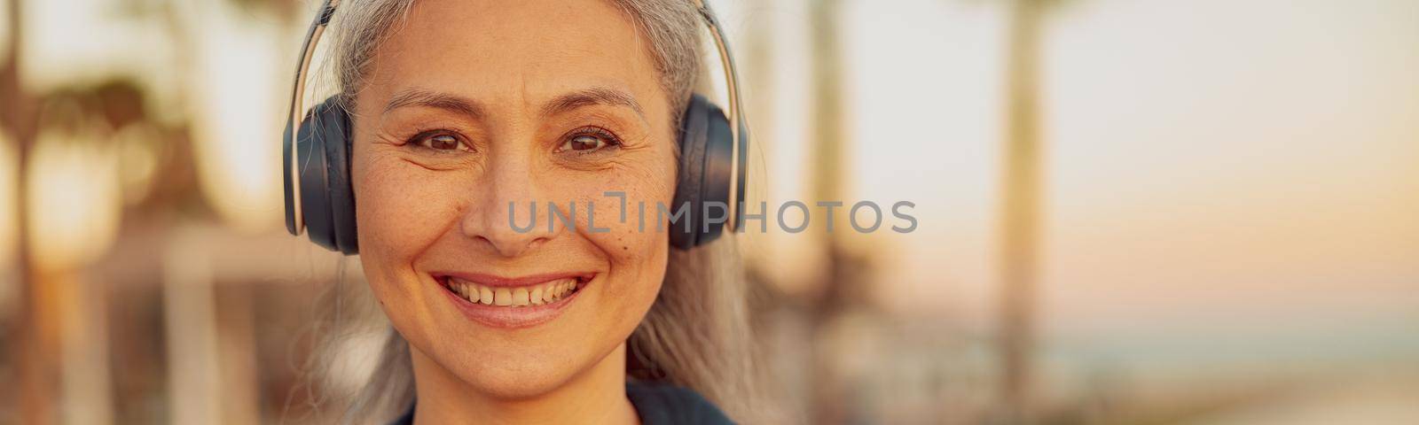 Close-up photo of Asian lady smiling at camera, wearing headphones and black hoodie on background of seafront with palm trees