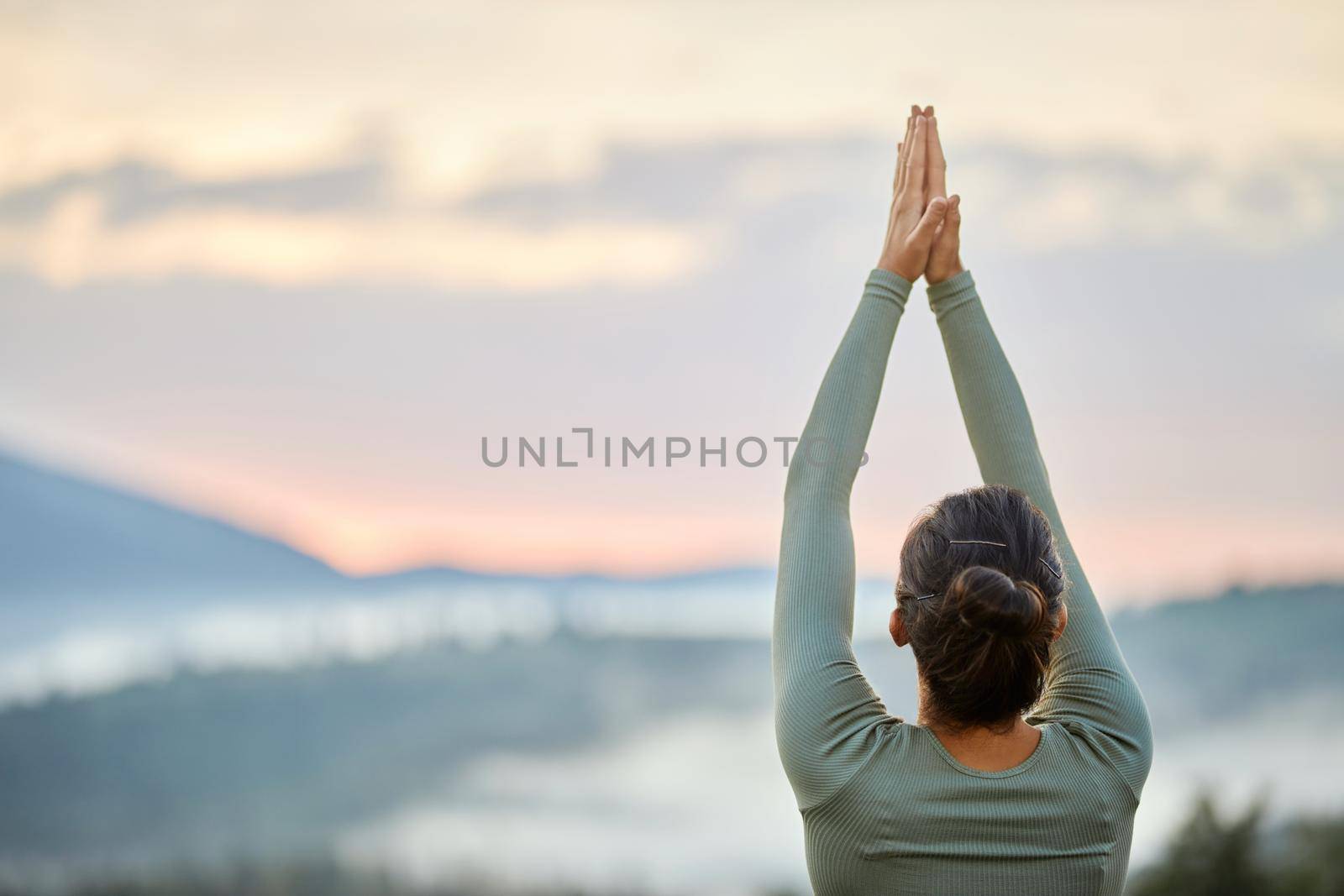 Back view of young girl in blue wear doing virabhadrasana on blur nature background. Crop of woman standing in warrior pose raising hands up folding together in cloudy mountains. Concept of balance.