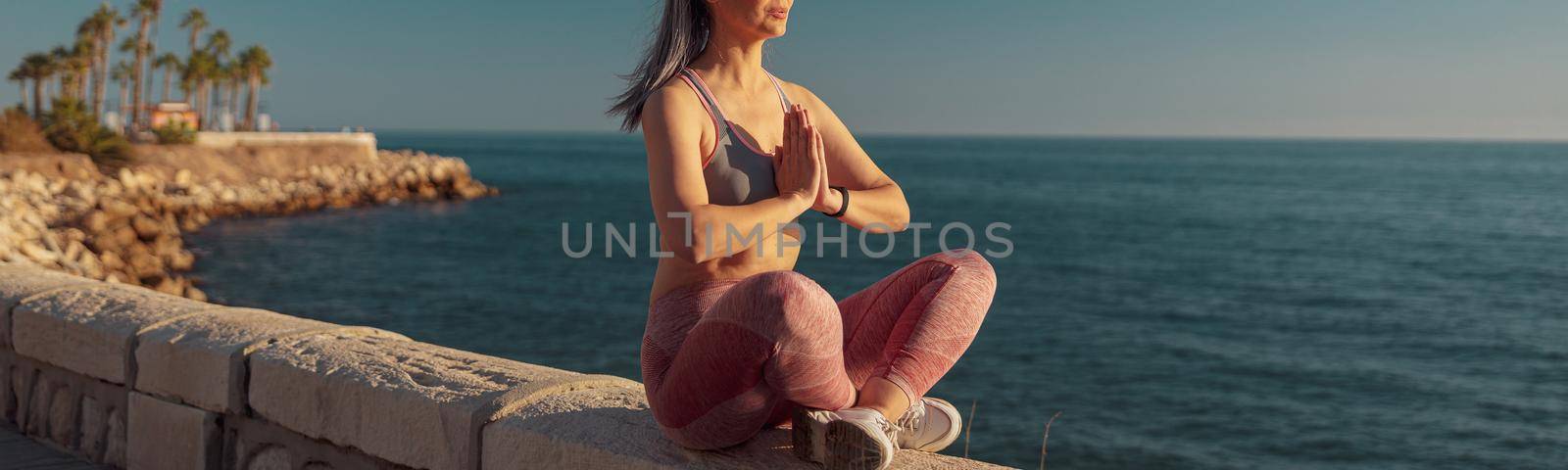 Sporty woman in sports clothes sitting by the beautiful seashore in lotus position and taining on warm day
