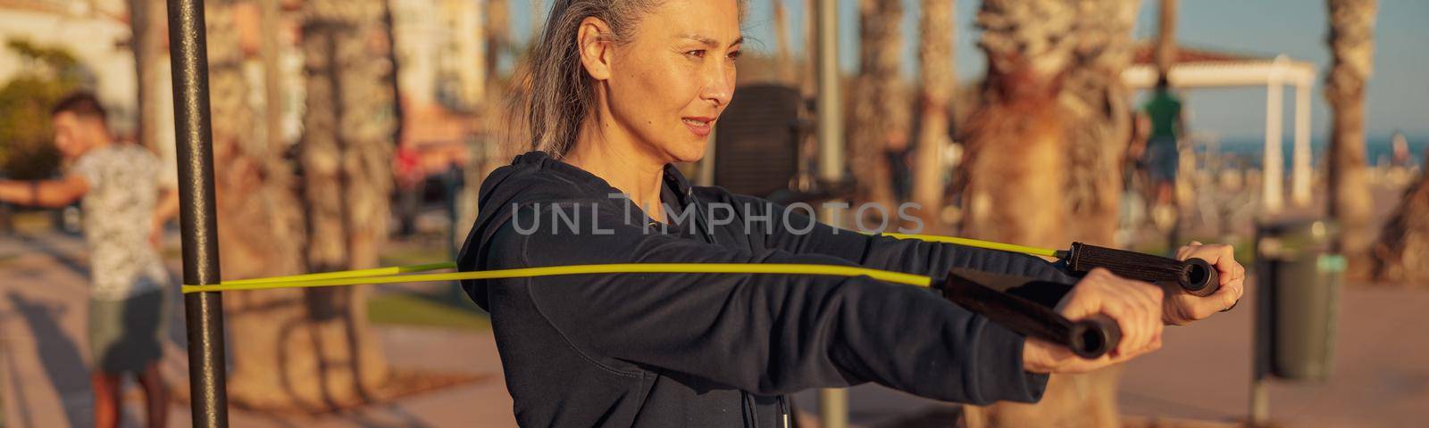 Focused sortswoman in black hoodie exercising using rubber bands, standing on sports site with arms extended forward