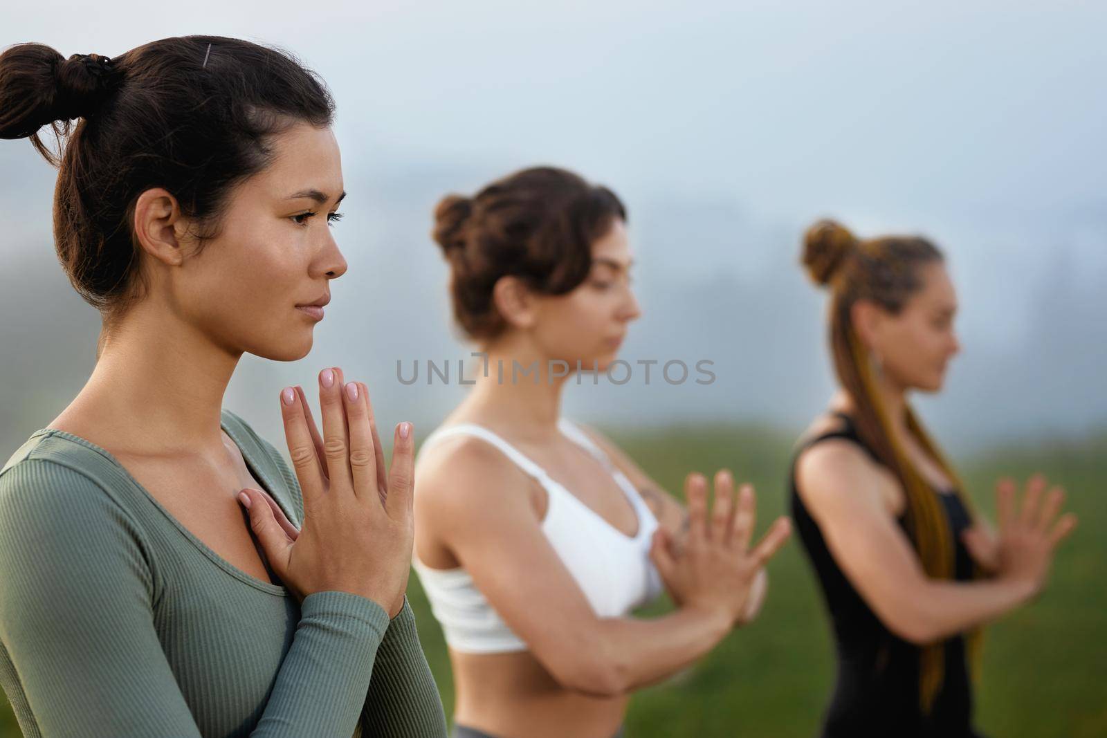 Crop of young brunette female near blurring girls putting palms together practicing yoga outdoors in morning. Close up of women standing in namaste poses. Concept of calmness and inner peace.