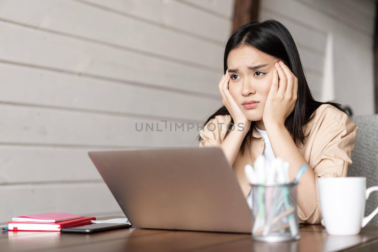 Upset and moody asian girl sits near laptop at home, looking disappointed, tired of studying remotely, bored of online classes.