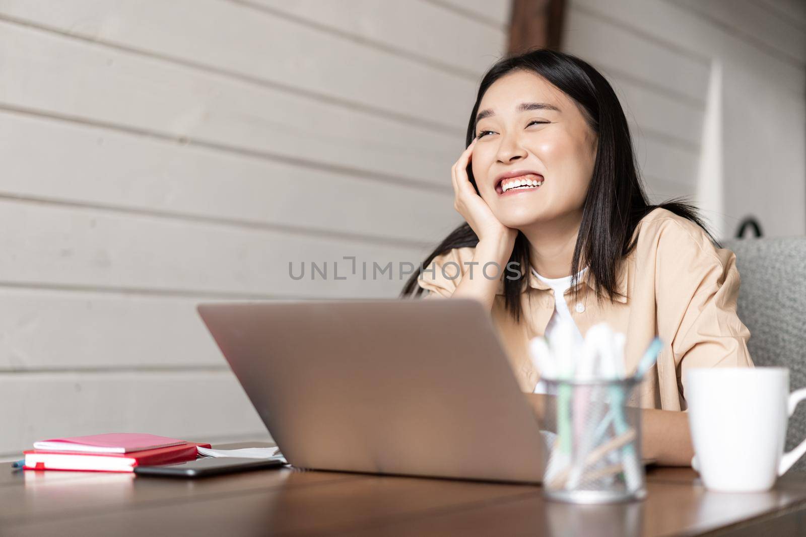 Asian girl student working on laptop, laughing and smiling, talking to someone, studying together from home, doing homework.