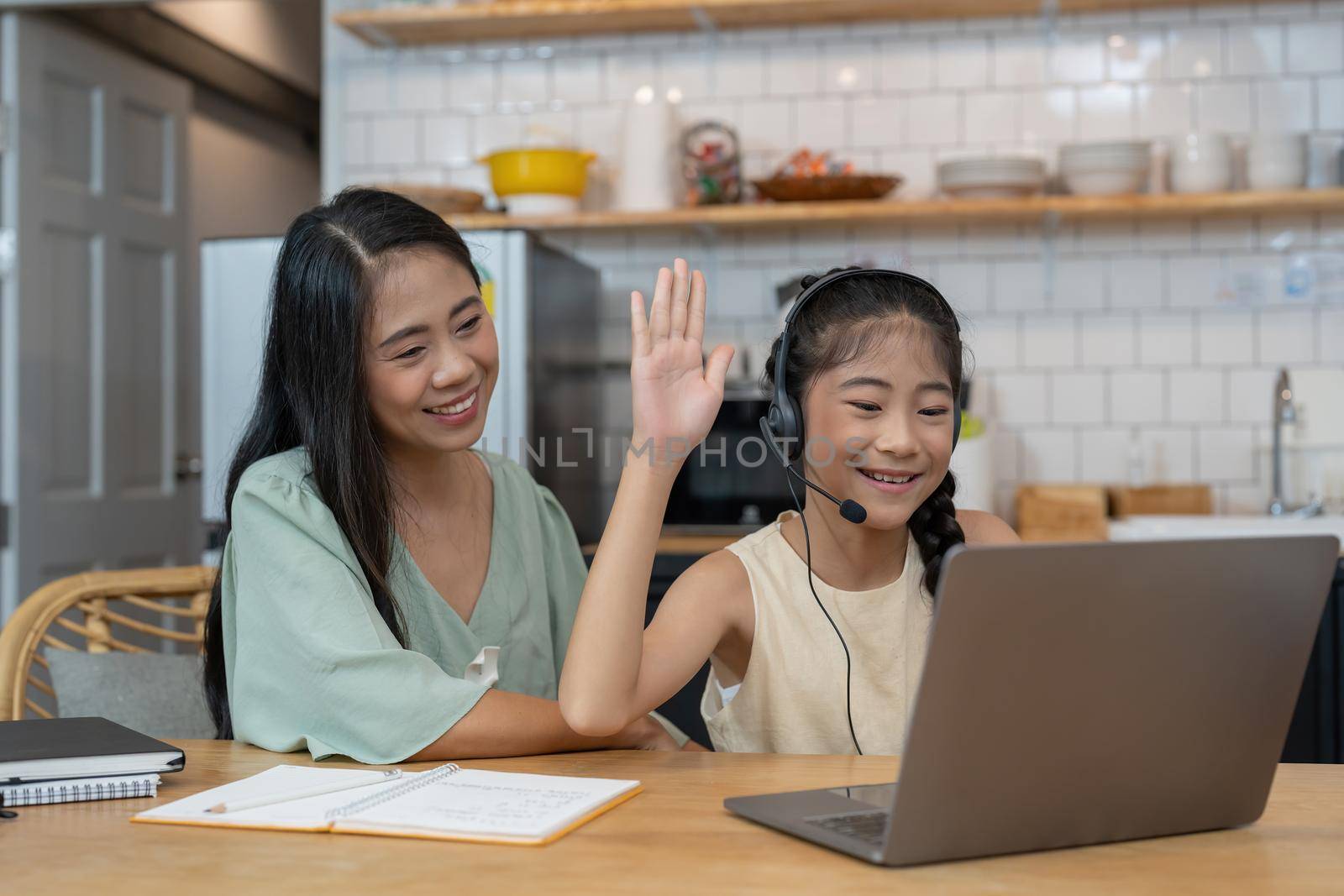 Happy asian mother and child sitting at kitchen table with colored pencils, attending virtual drawing class via video call, smiling and waving hello at laptop computer screen to greet online teacher by nateemee