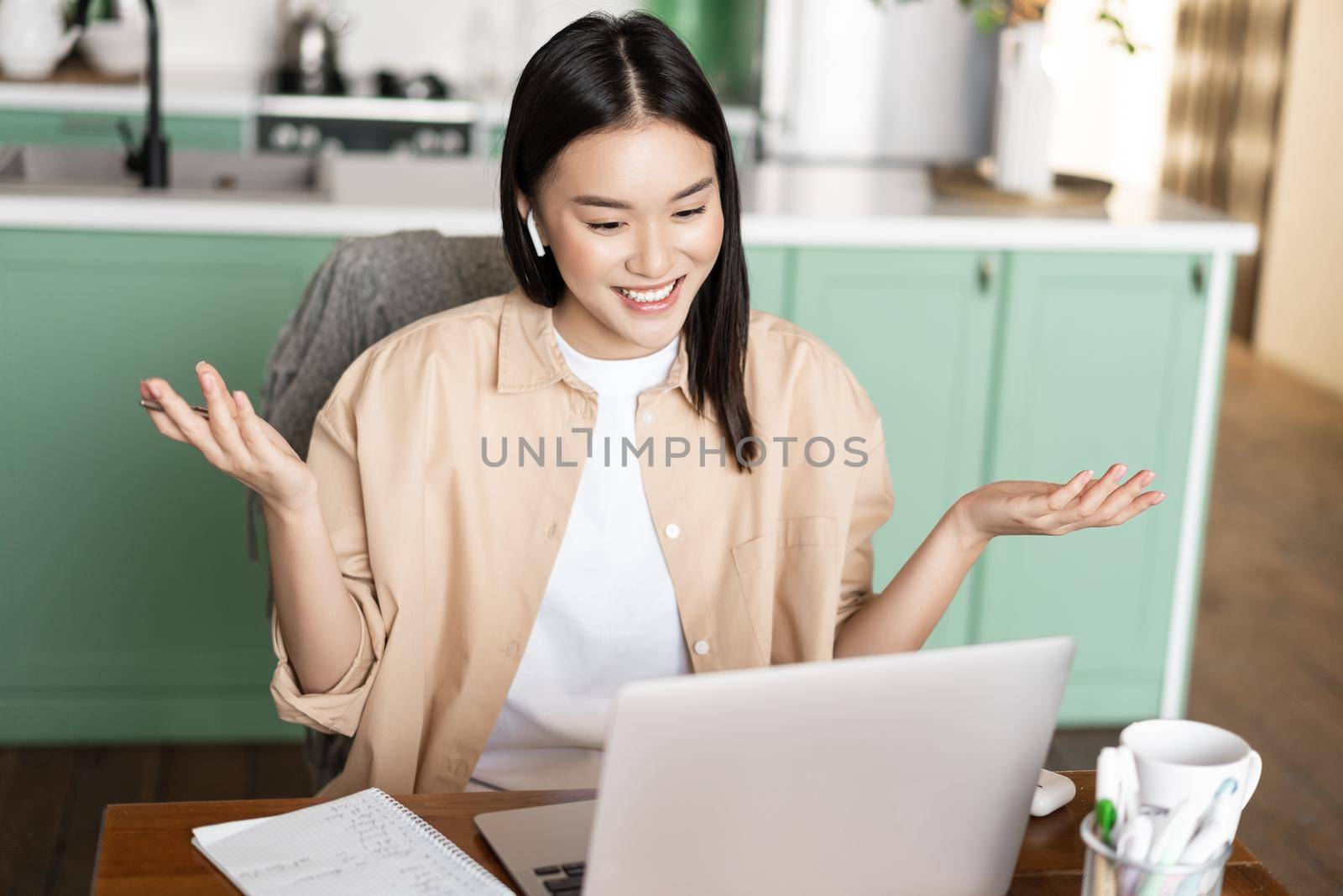 Asian woman tutor having online classes from home, working via laptop, having video call conference, wearing headphones, sitting at table surrounded by study material.