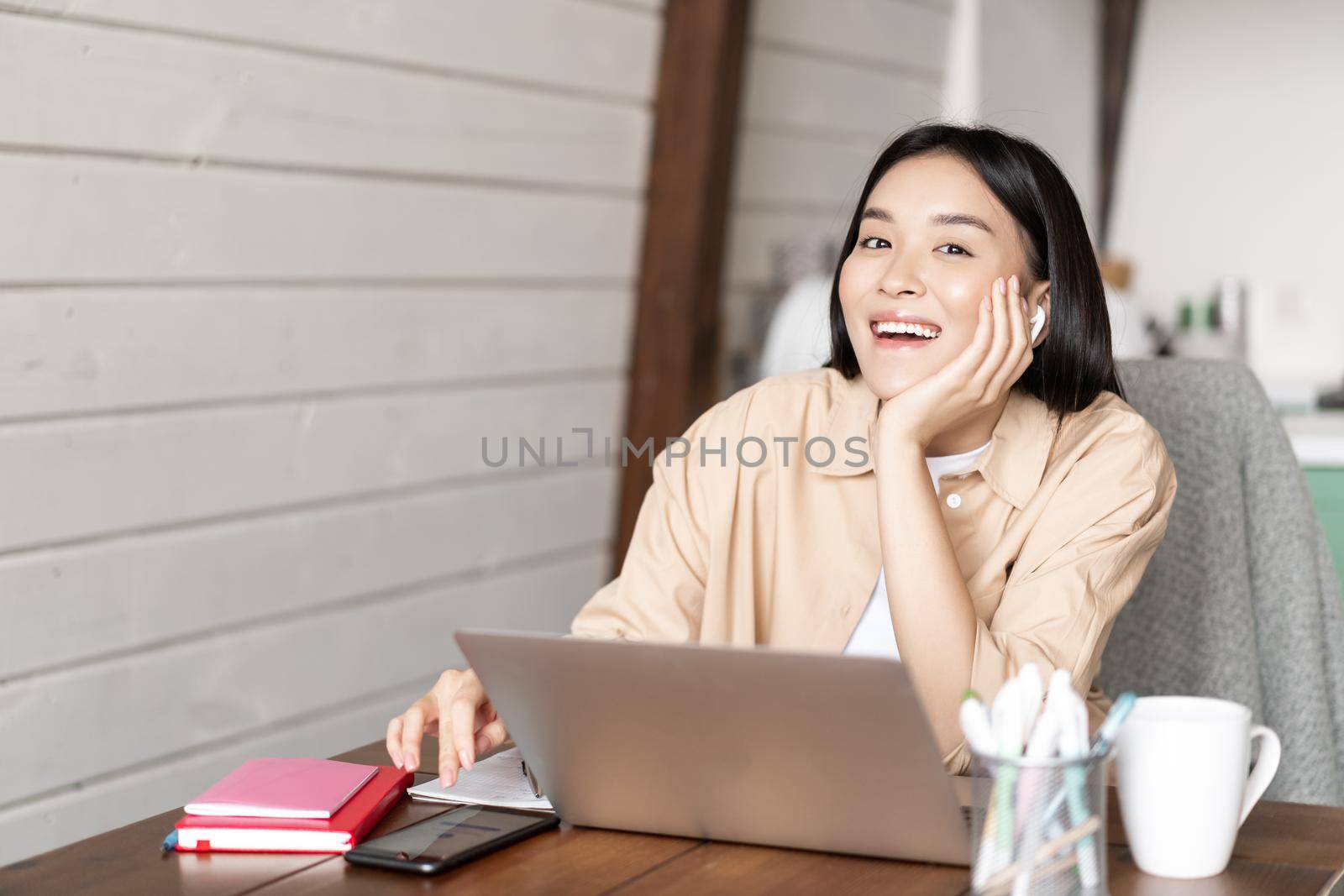Happy asian girl listening online webinar, attend internet course classes on laptop, wearing wireless earphones and smiling, working from home.