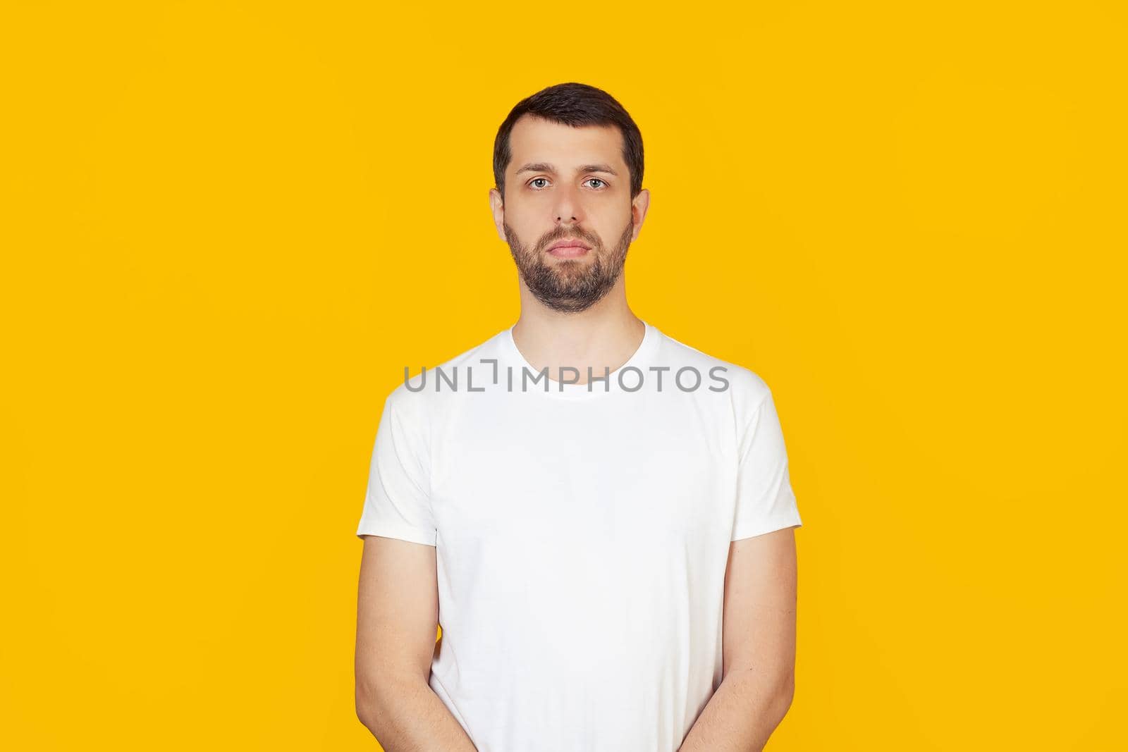 Young man with a beard in a white t-shirt with a serious expression on his face. Simple and natural looking into the camera. Stands on isolated yellow background.