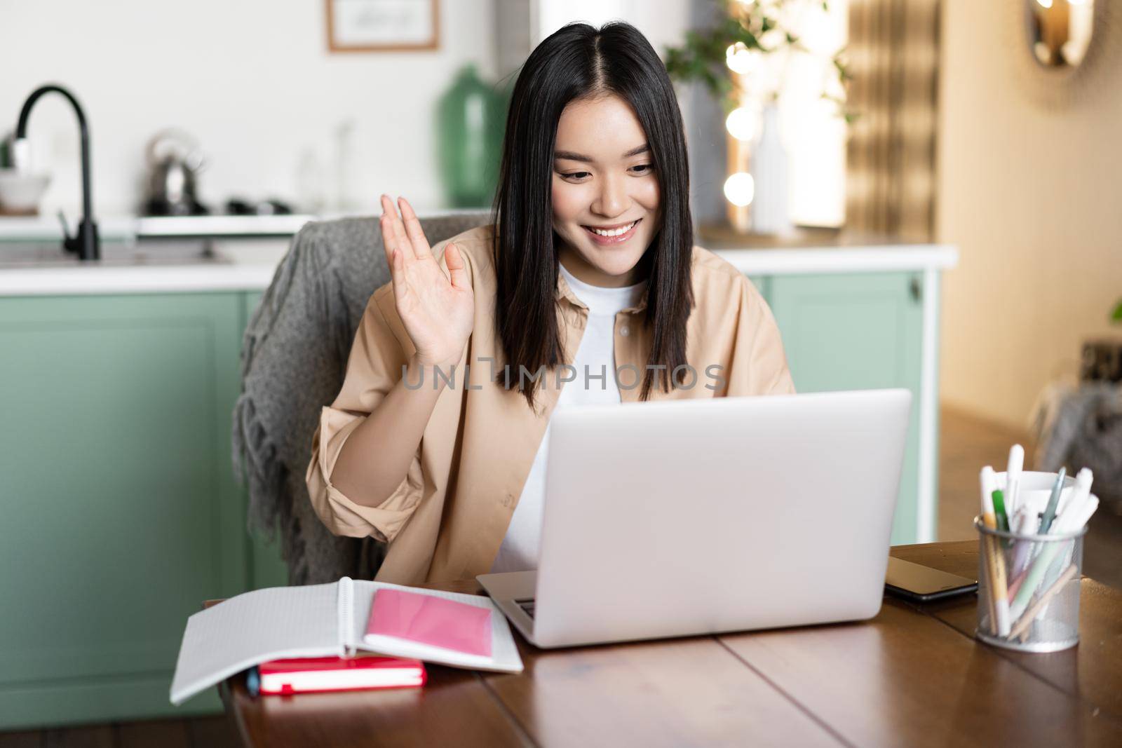 Image of asian girl on video call, waving hand at her video chat on laptop and smiling. Student or teacher greeting people during remote class online.