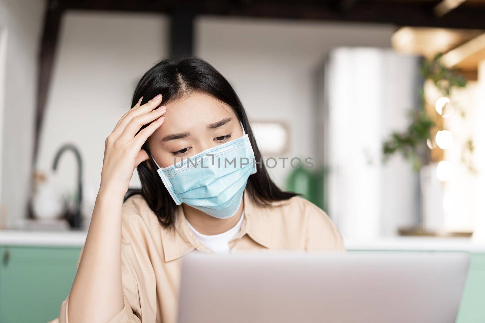 Tired asian girl in medical face mask listening webinar, watching smth on laptop screen with troubled face, doing homework.