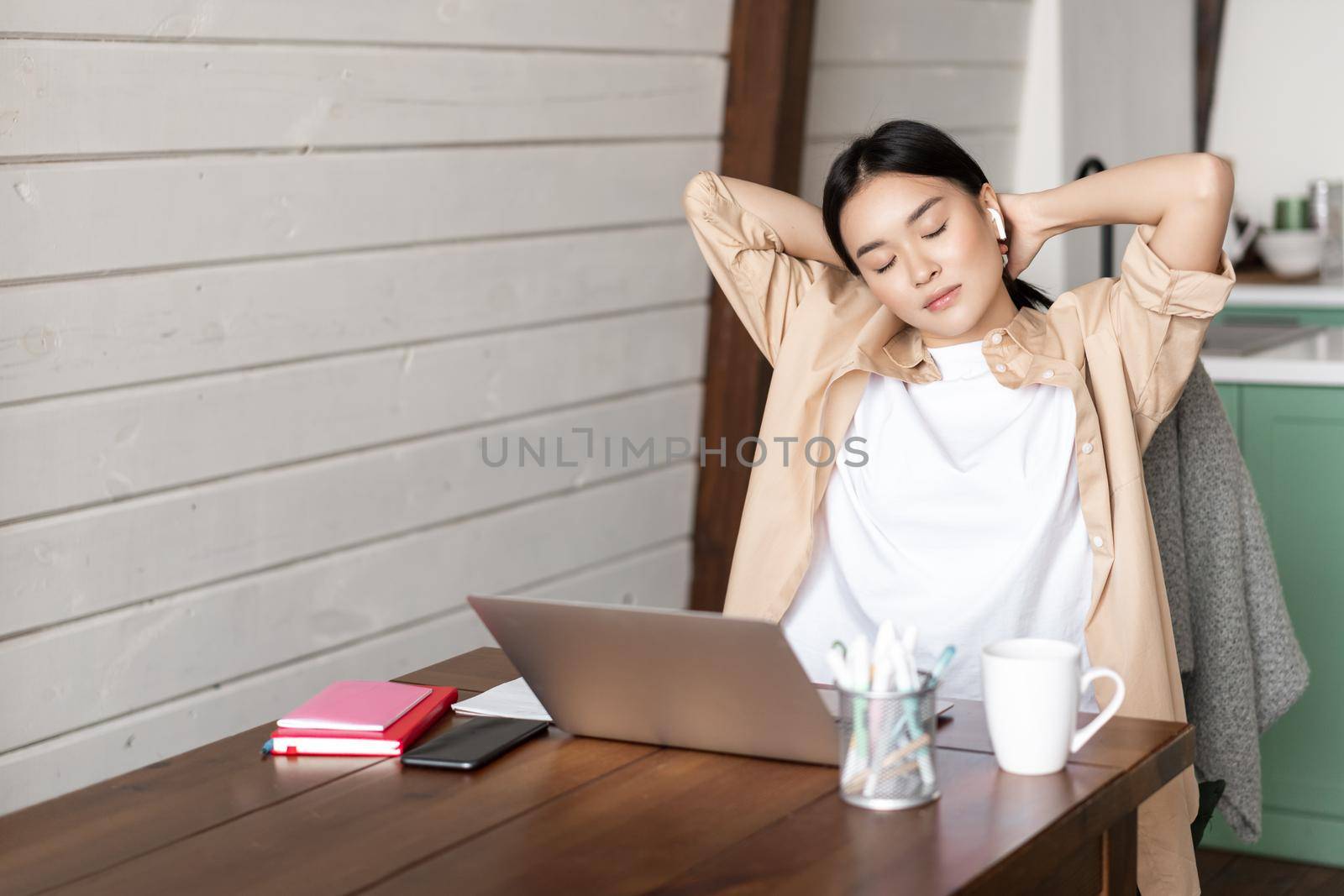 Young asian woman takes break from work, stretching arms tired of working on laptop, finishes homework, studies remotely from home in kitchen by Benzoix