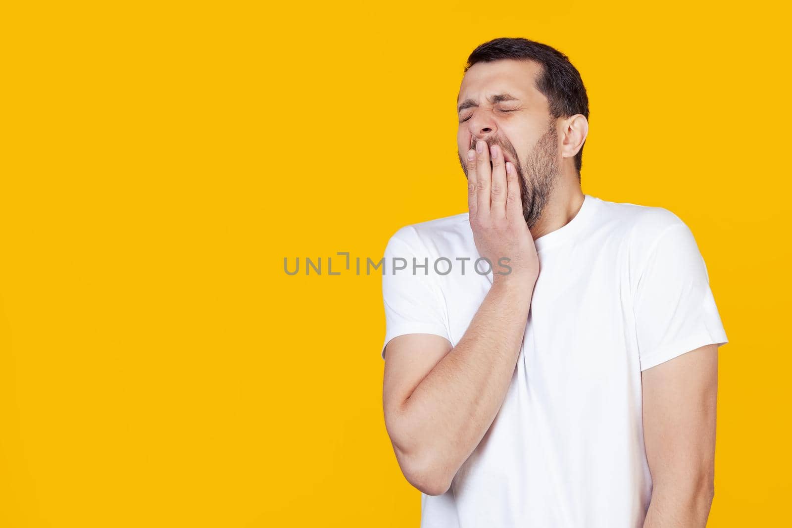 A modern young man with a beard in a white T-shirt yawns boredly against a yellow background, tired of covering his mouth with his hand. Restlessness and sleepiness