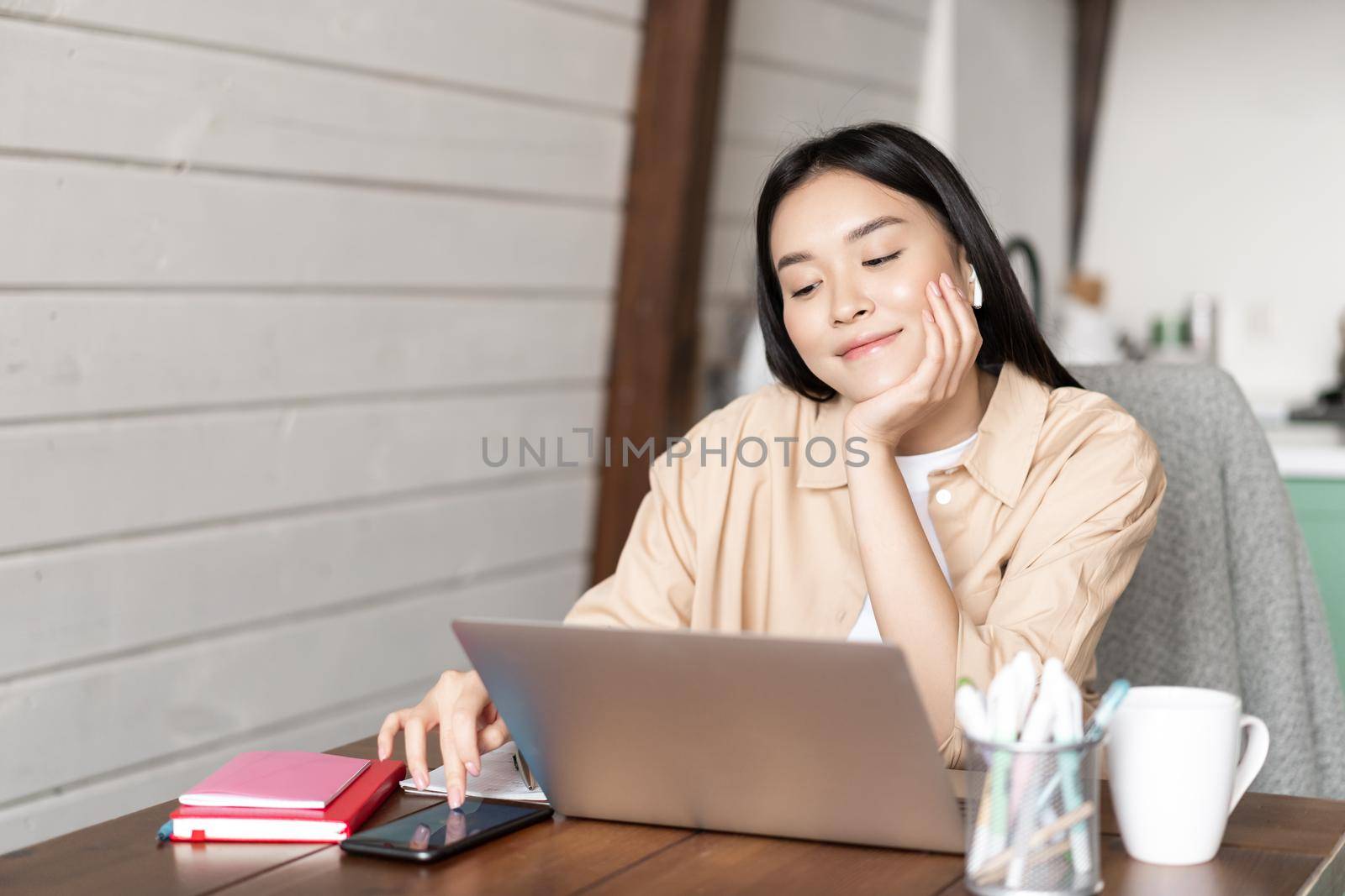 Smiling asian girl student, woman sitting with laptop at home kitchen, browing smartphone social media and resting during online classes break, waiting for webinar.