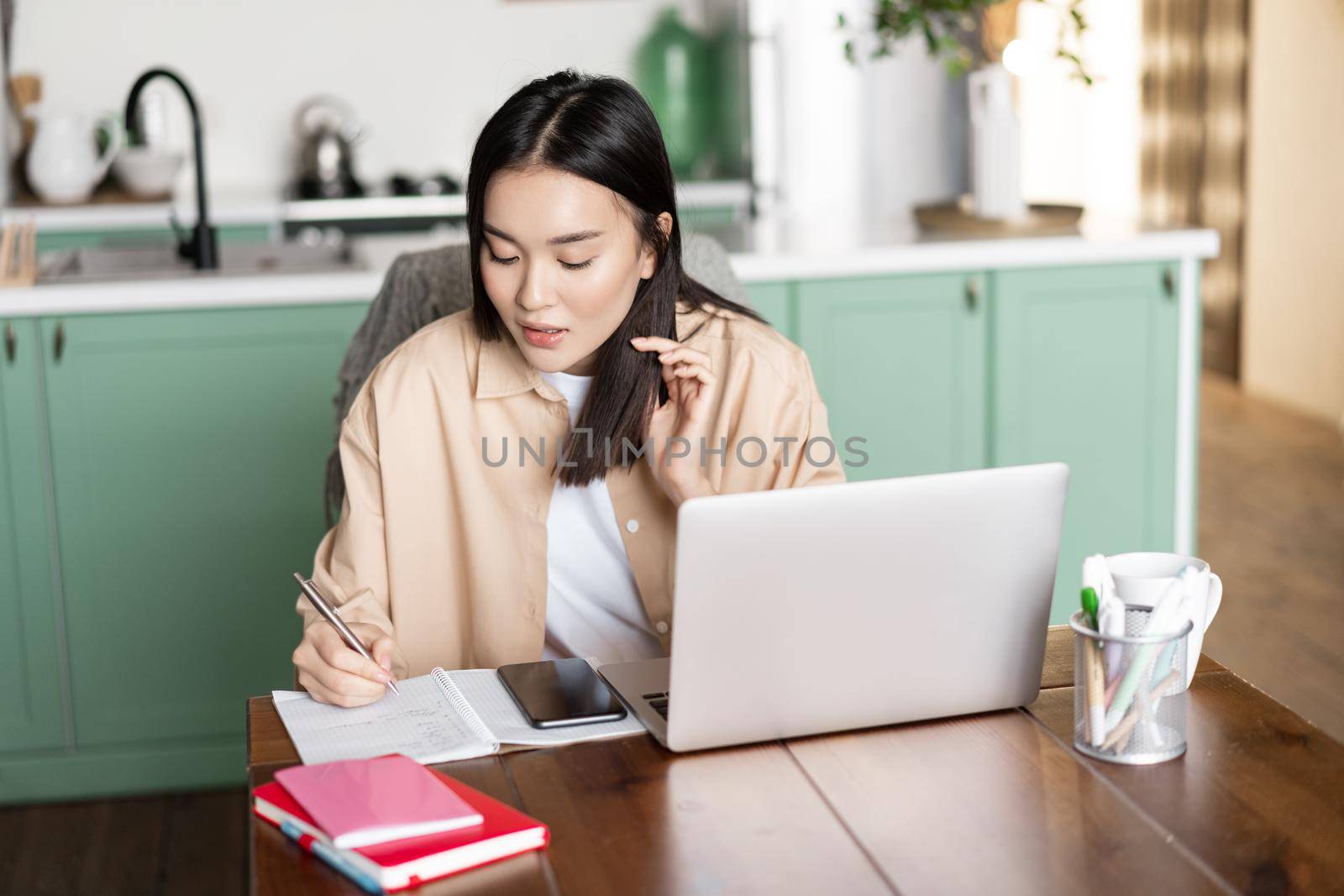 Asian girl student doing homework at home. Young woman taking notes, working on laptop and writing down info.