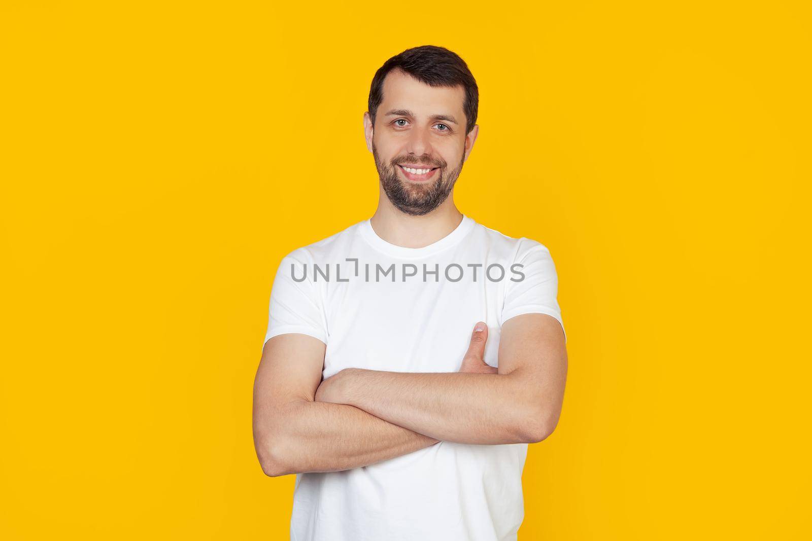 Young man with beard in white t-shirt happy face smiling with crossed arms looking at the camera. Positive man stands on isolated yellow background.