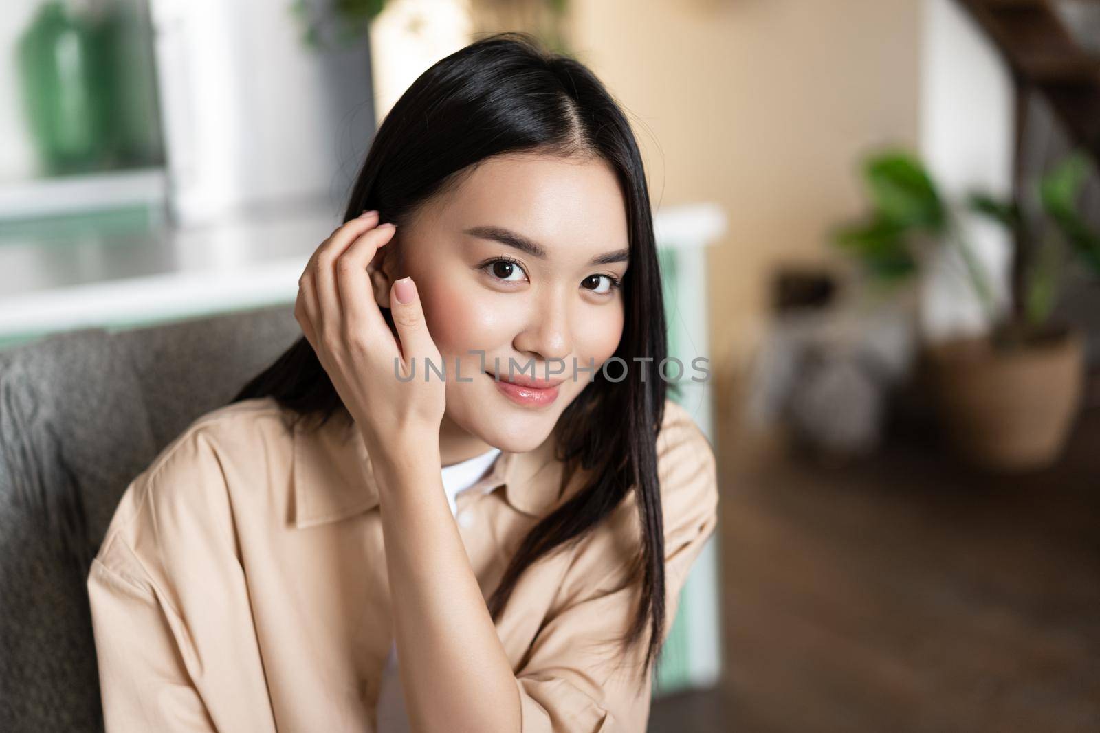 Portrait of asian woman smiling, sitting at home. GIrl touches her hair and looks cute at camera by Benzoix