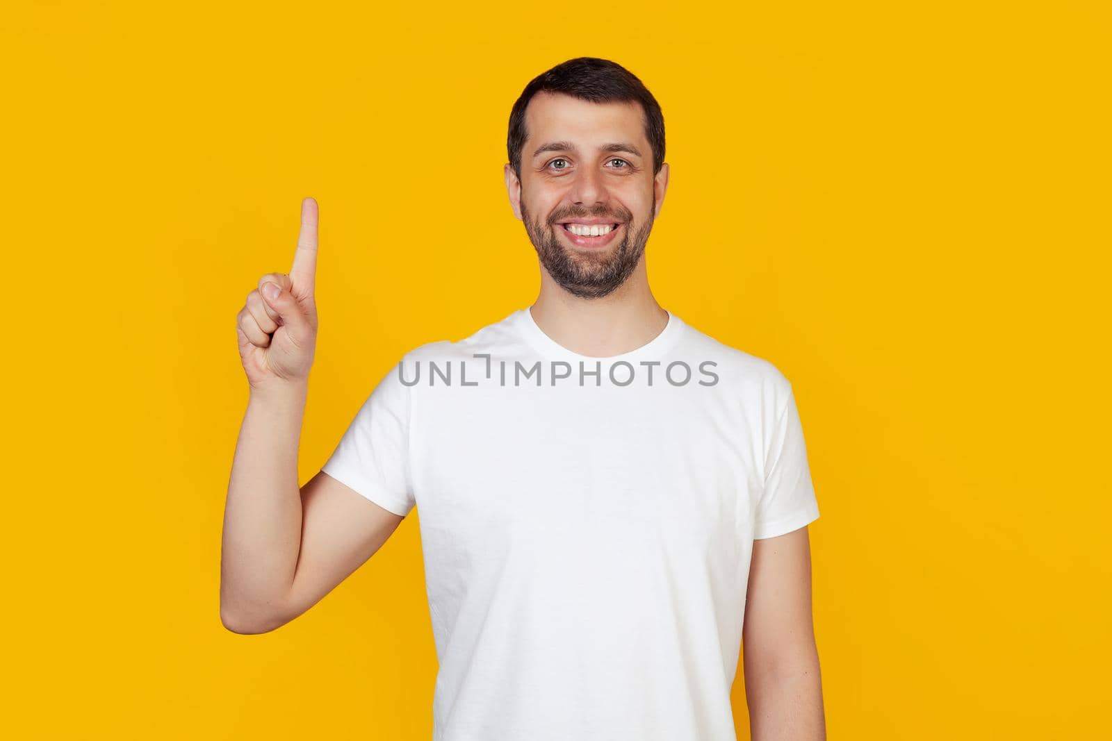 Modern young man with a beard in a white tank top shows number one with fingers on hand, smiling confidently and happily, looking at the camera. The man shows one finger. Number 1, yellow background